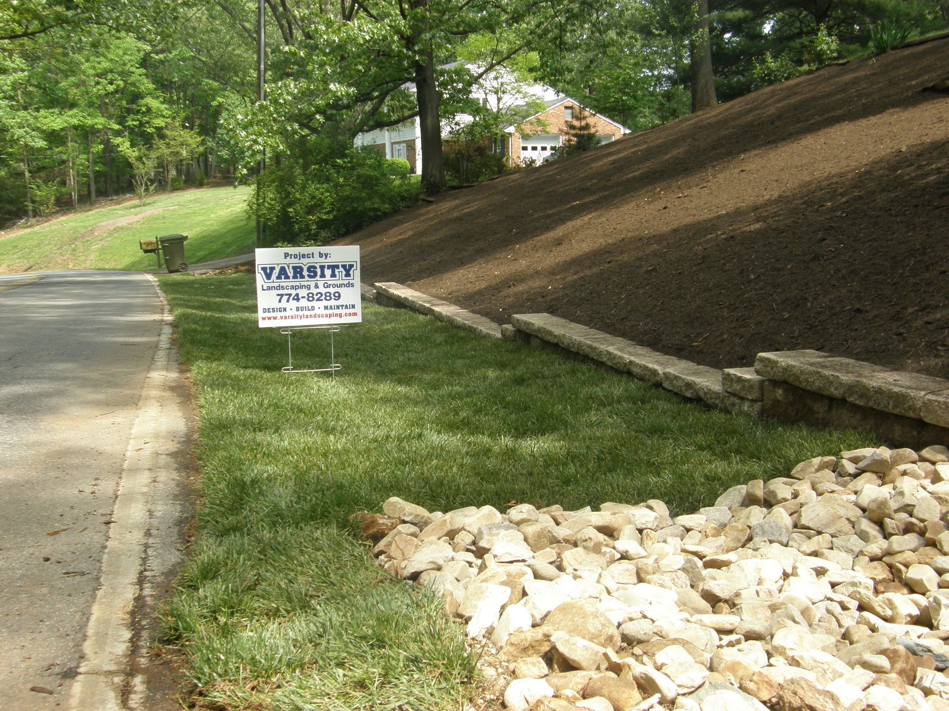 Roadside warning sign beside a sloped dirt bank and rock pile next to a paved path in a wooded area