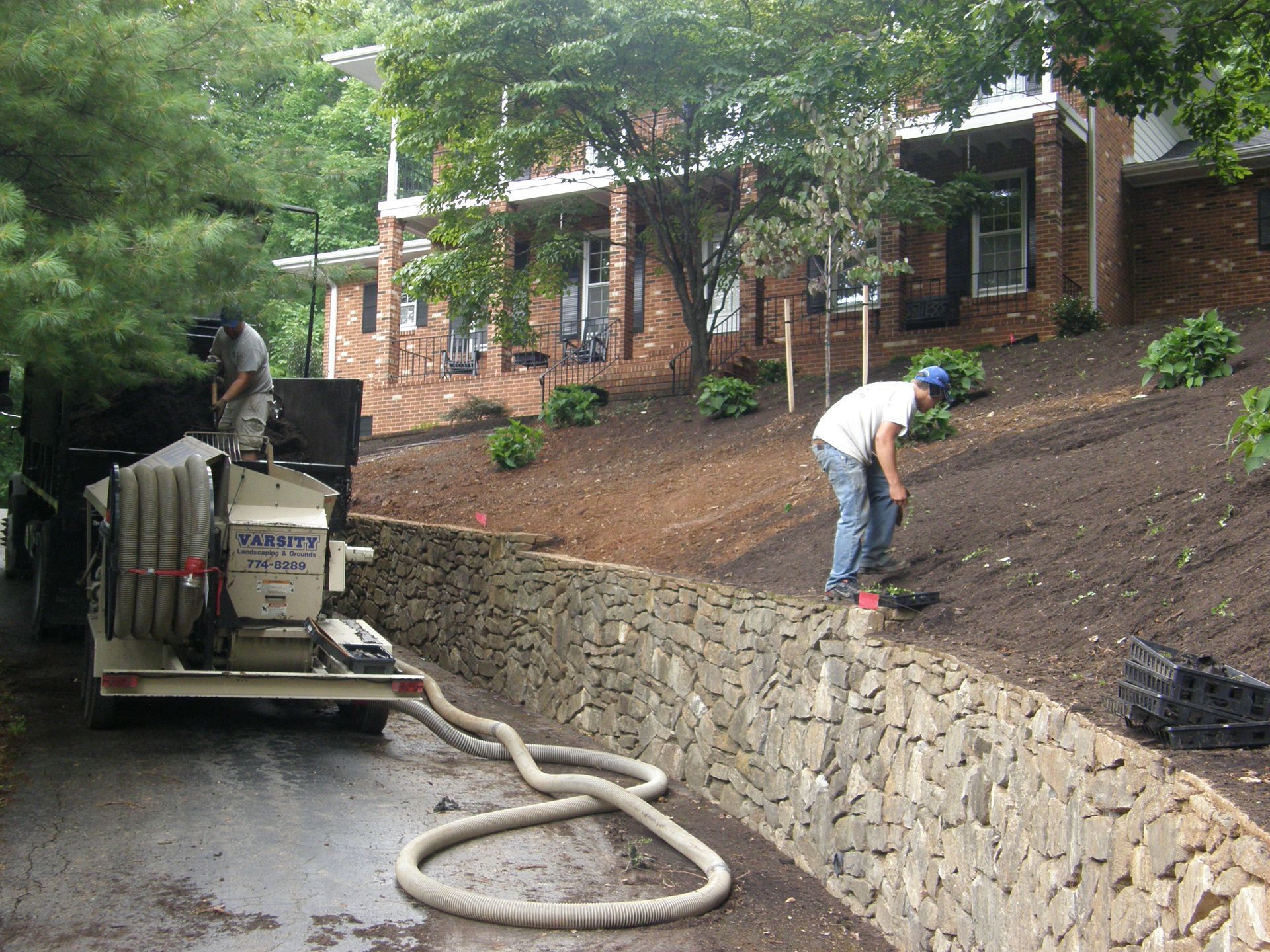 Workers installing landscaping on a hillside beside a stone retaining wall, with a truck and hose in the foreground