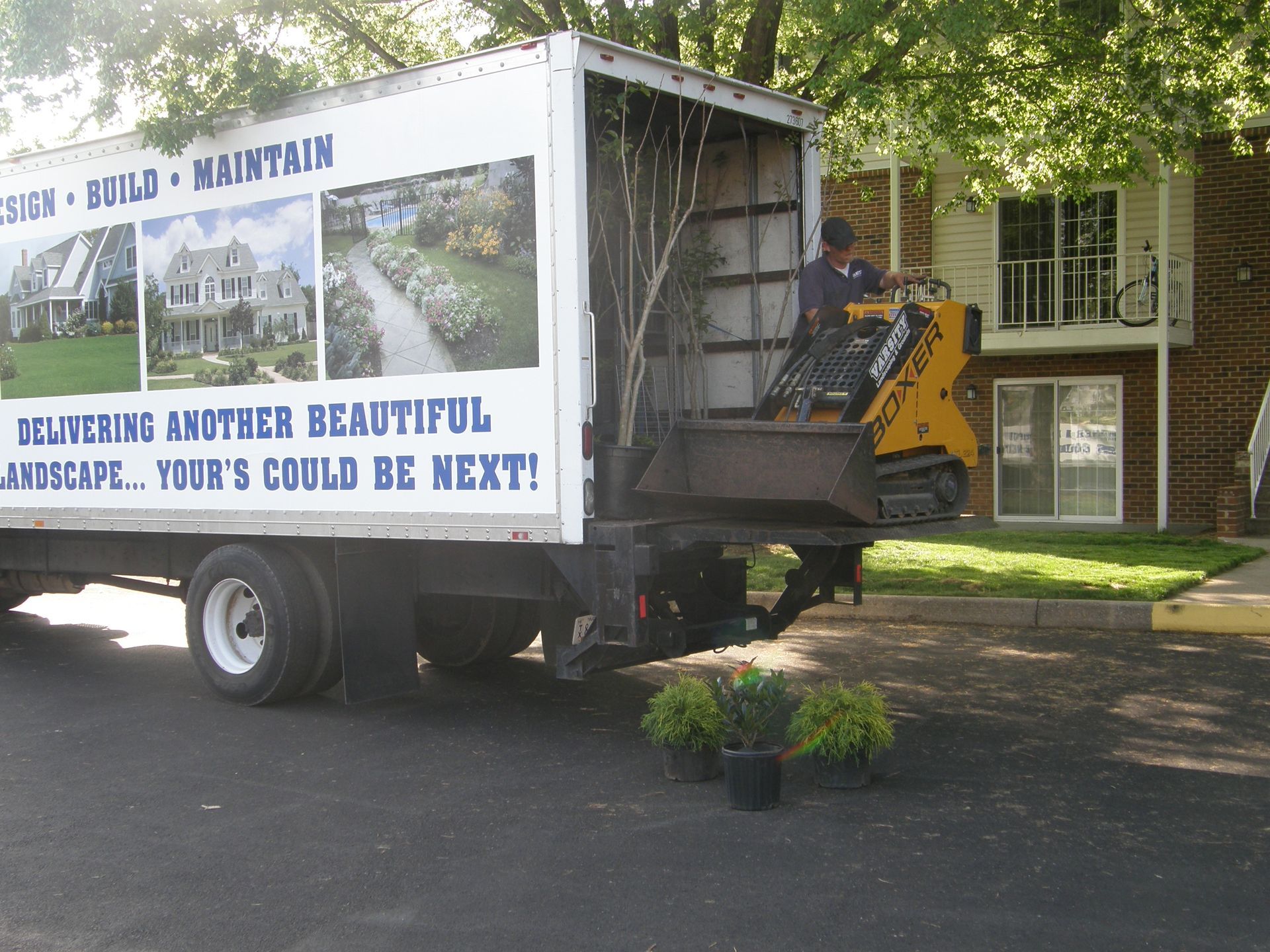 Moving truck with a lift parked by a house, carrying plants and a yard sign about replacing another beautiful landscape.