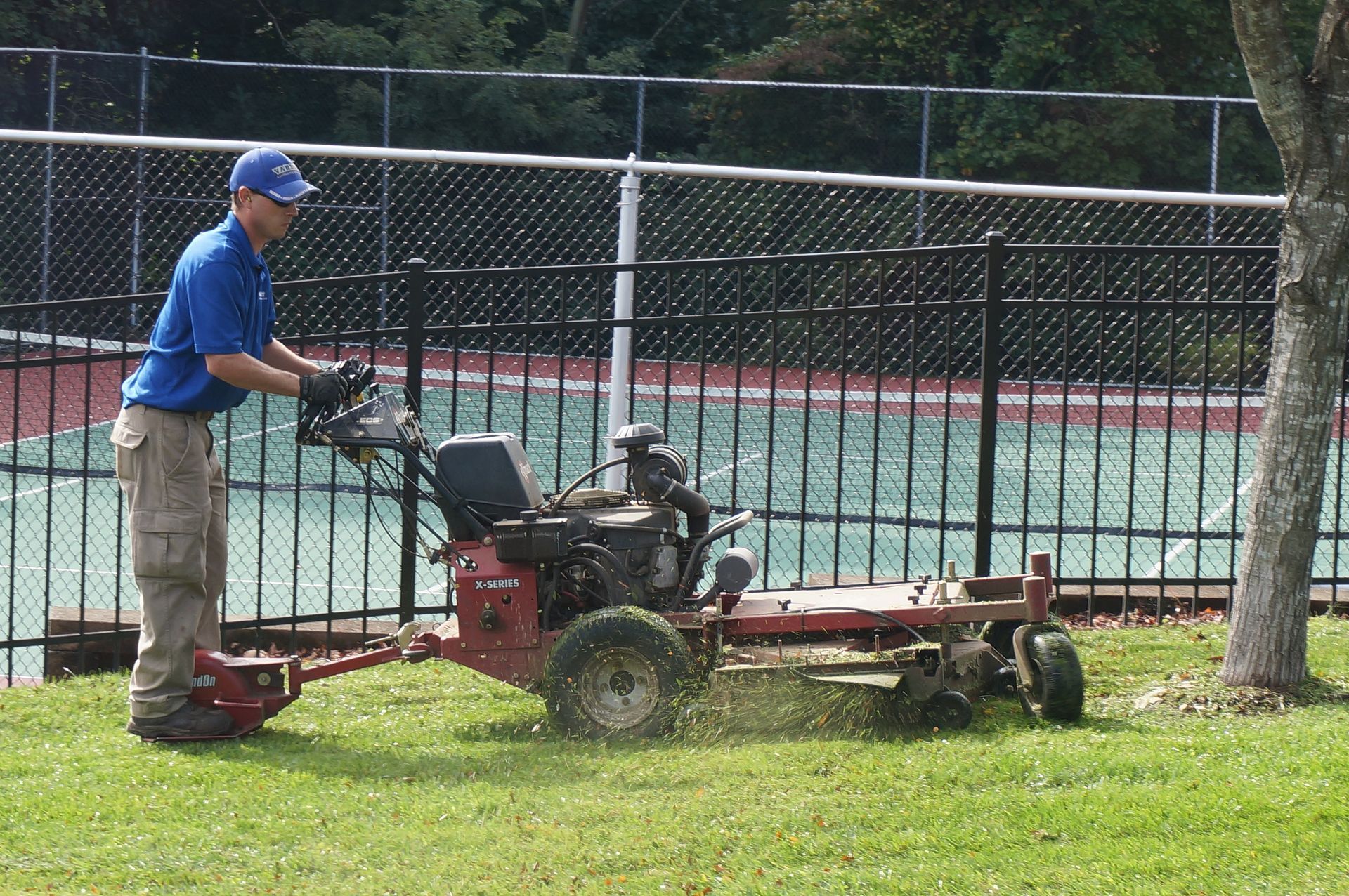 Worker mowing grass with a riding mower beside a fence and tennis court