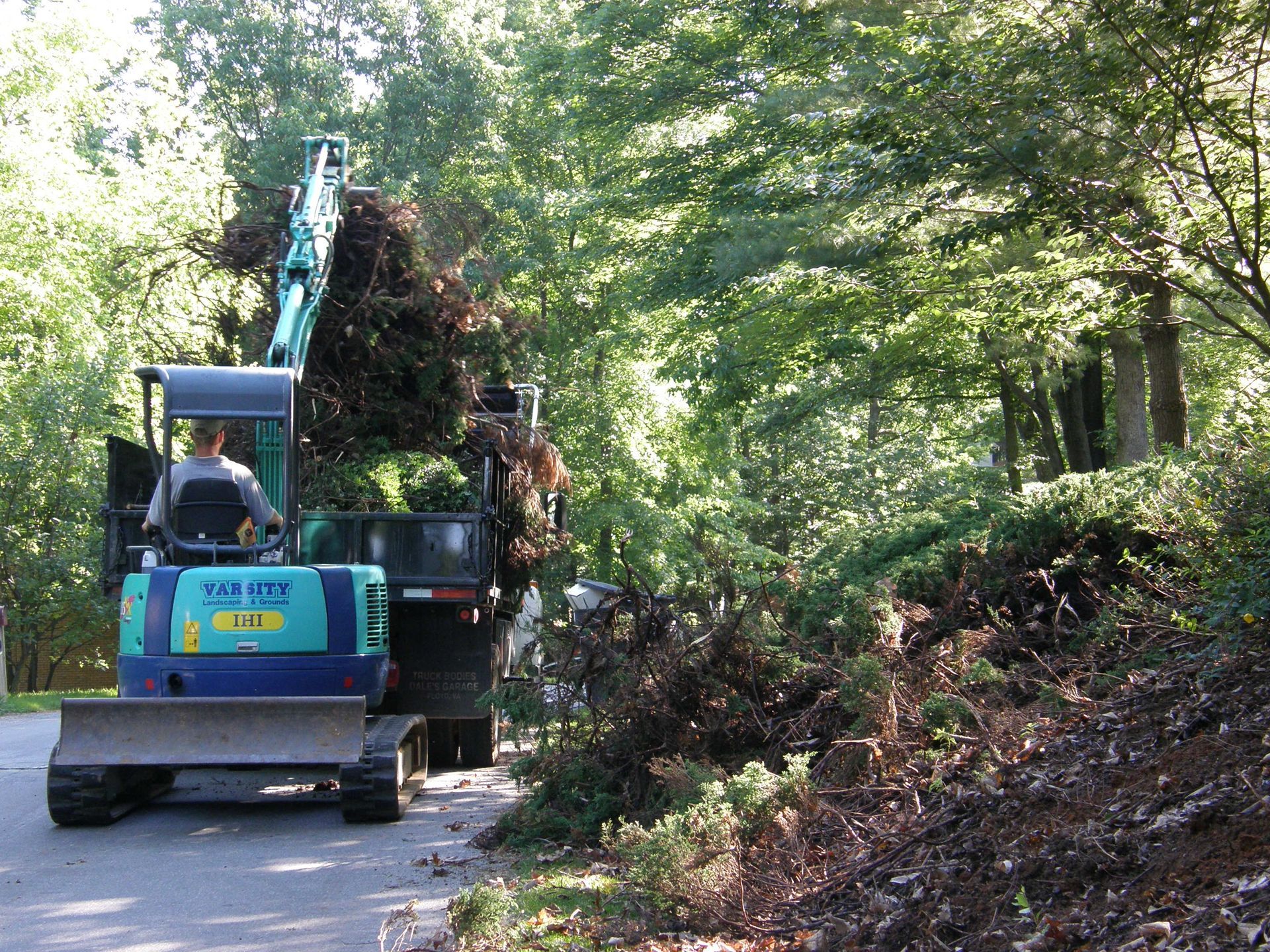 Blue excavator clearing brush along a wooded road with piled branches on the right.