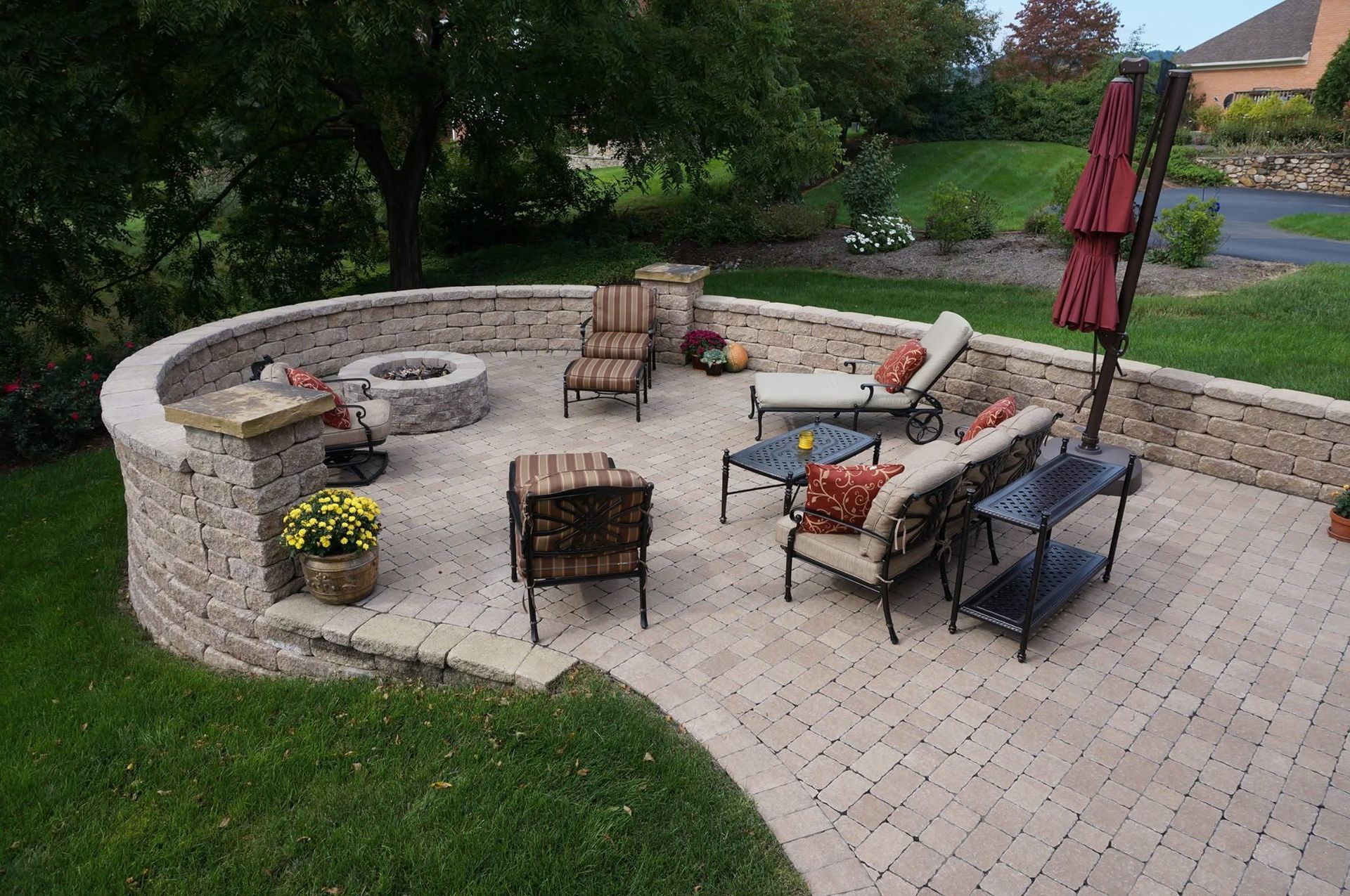 Outdoor stone patio with wicker chairs, round fire pit, red umbrella, and potted flowers in a backyard garden