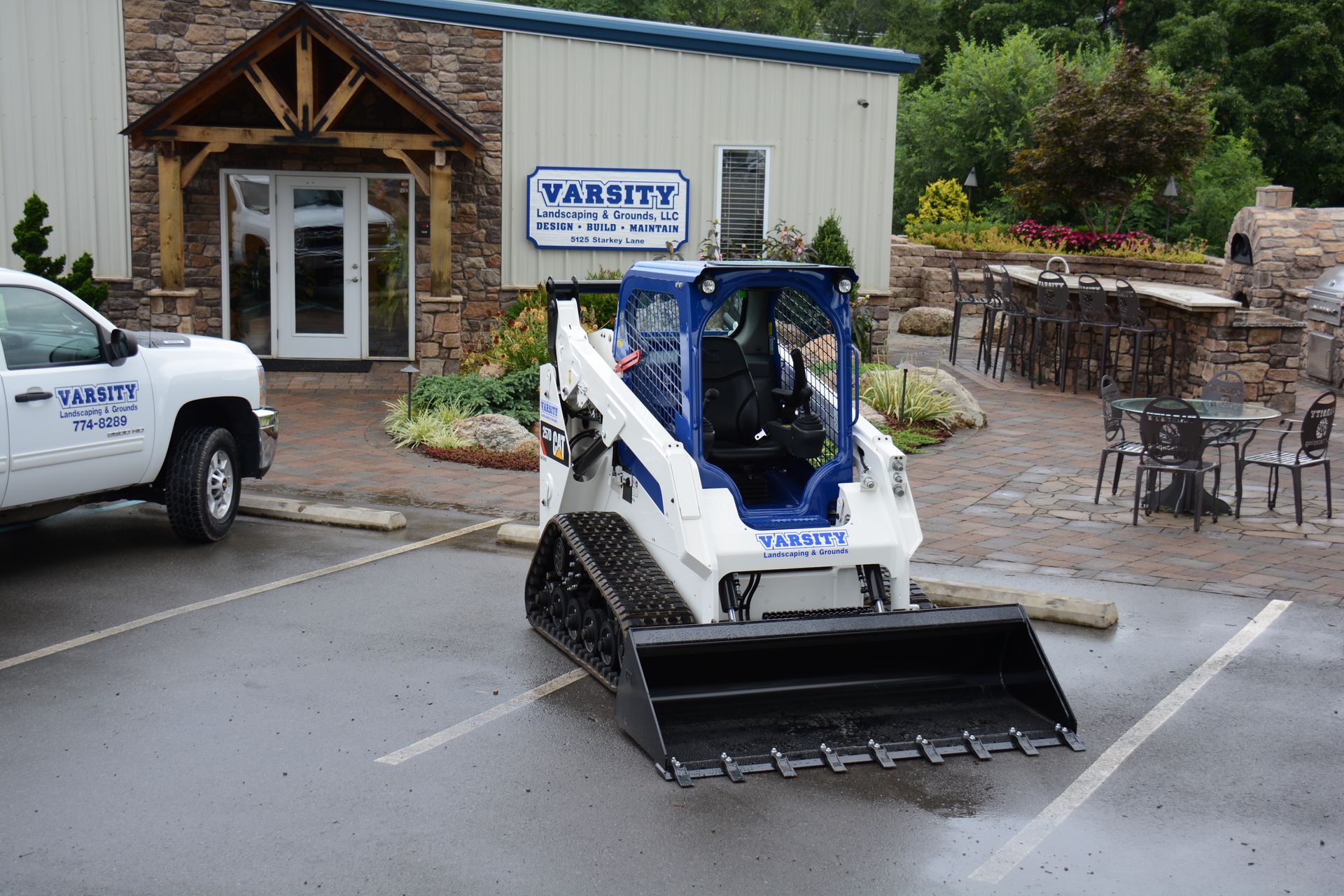 White skid-steer loader with snow plow parked outside a building, with a white truck nearby.