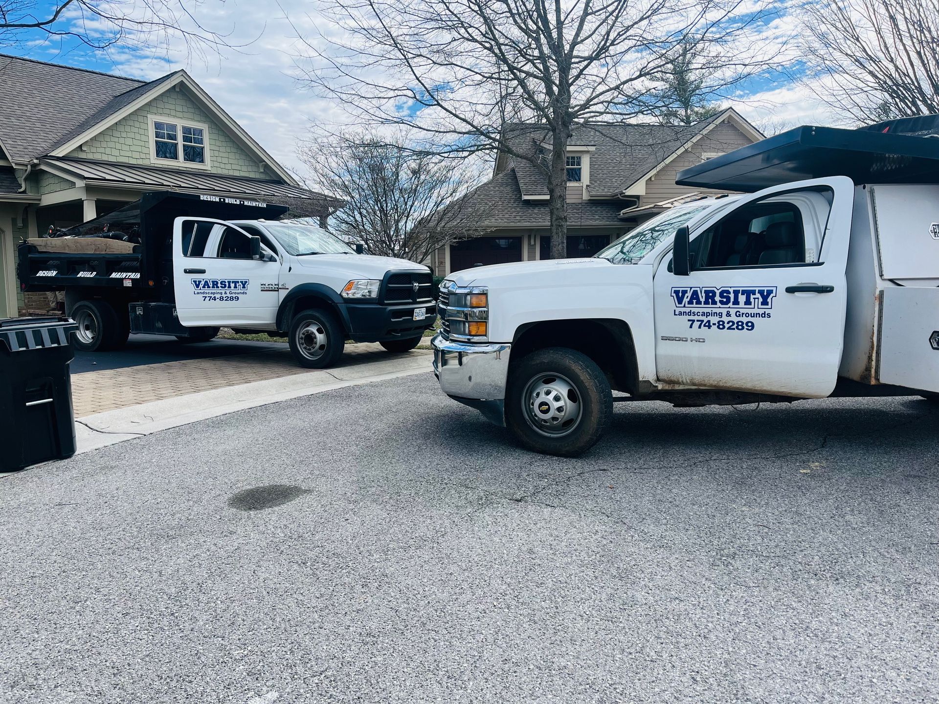 Two white utility trucks parked in a driveway by a house, one with open passenger door.