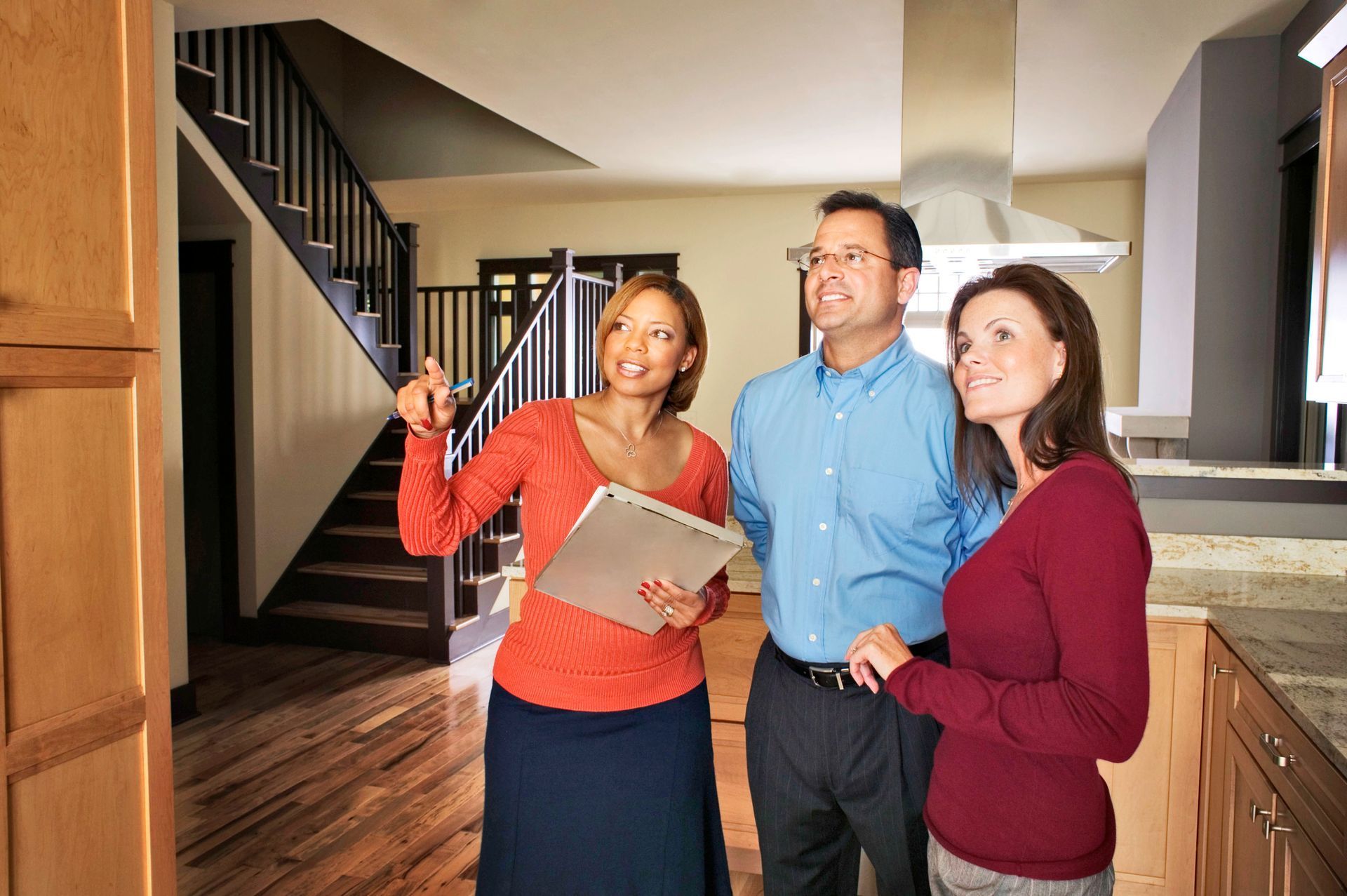 Real estate agent showing a couple a home. Agent points, couple smiles, interior kitchen and staircase visible.