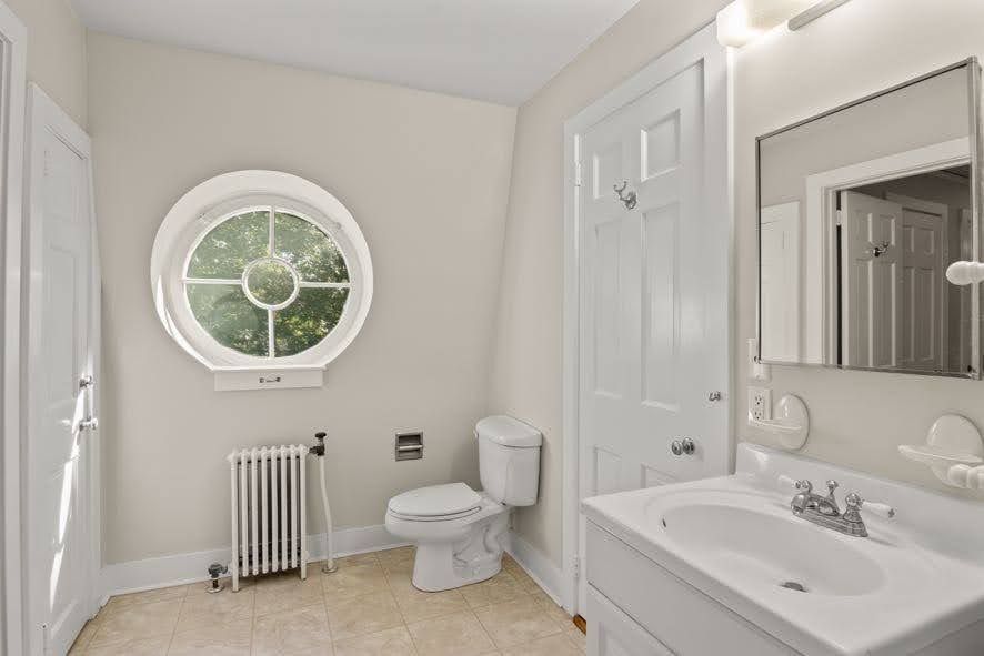 A bathroom with a toilet , sink , mirror and window.