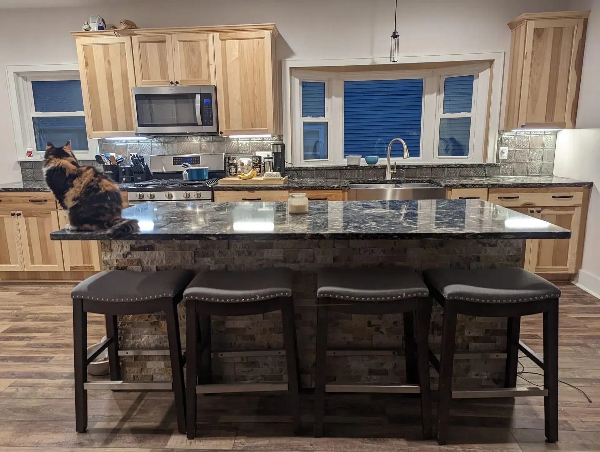 Kitchen with a cat on an island. Light wood cabinets, granite countertops, and dark stools.