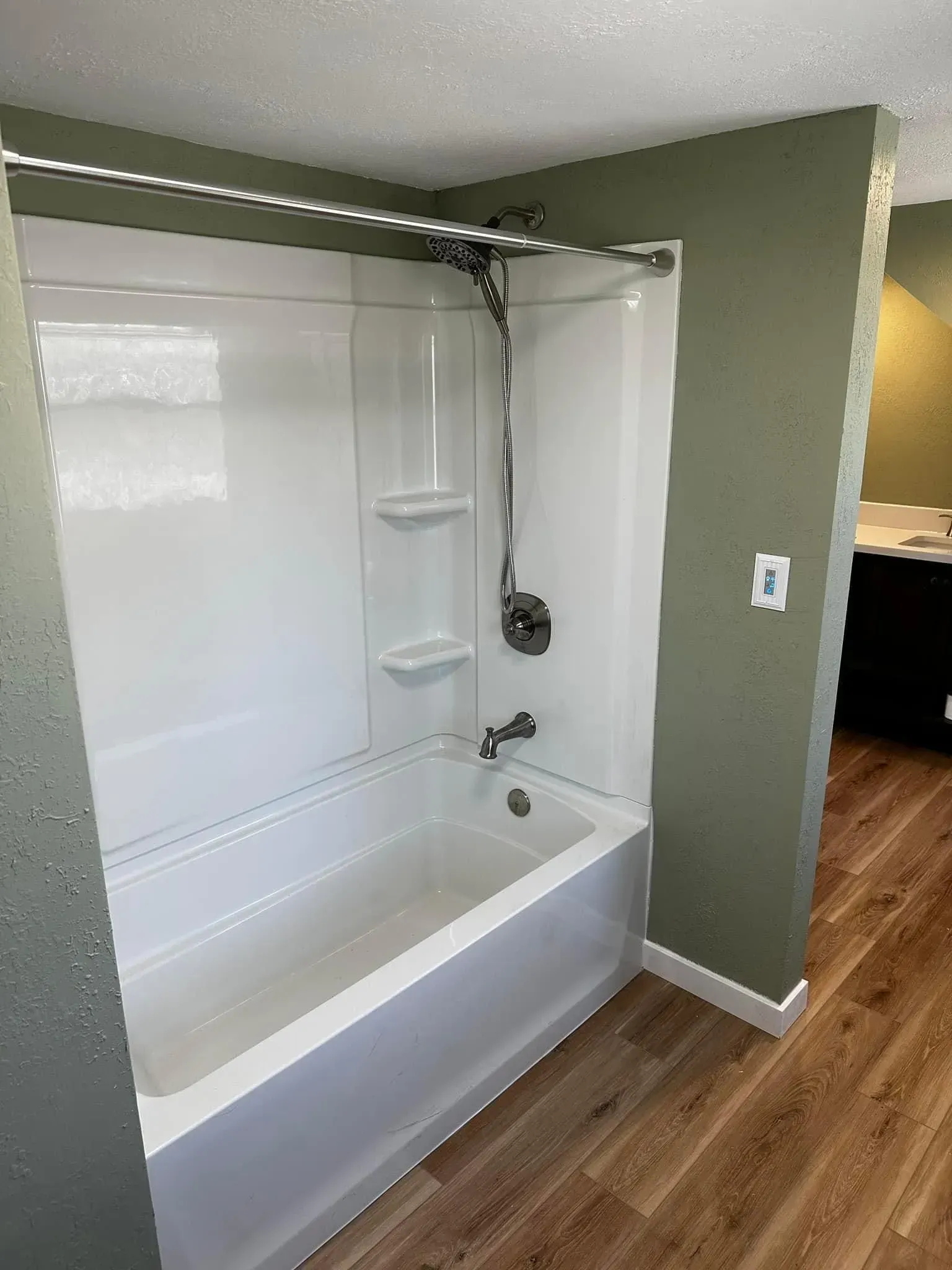 White bathtub and shower combo with a green wall, in a bathroom with wood-look flooring.