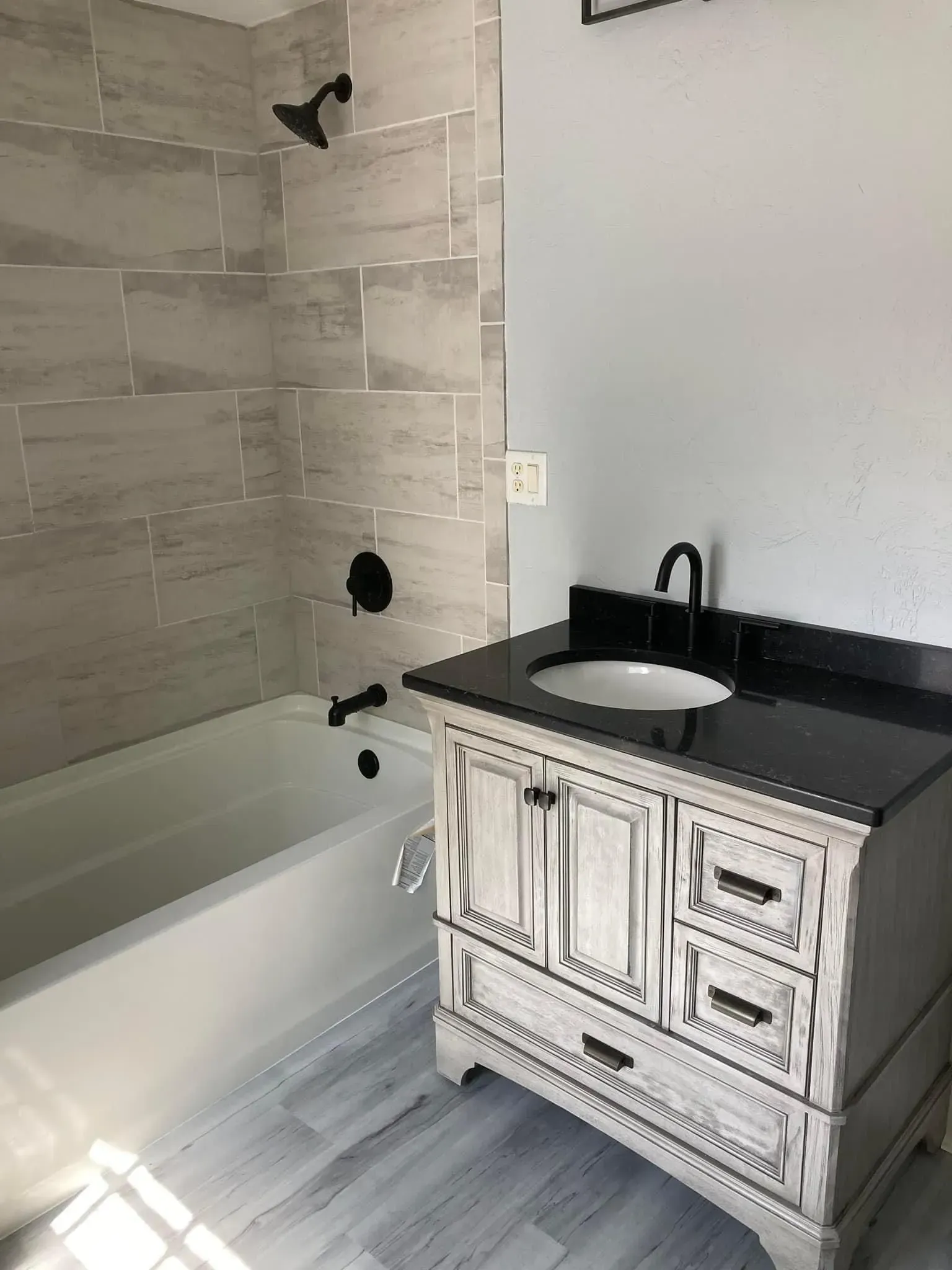 Bathroom with white tub, light gray tile shower, distressed white vanity with black countertop, and black fixtures.