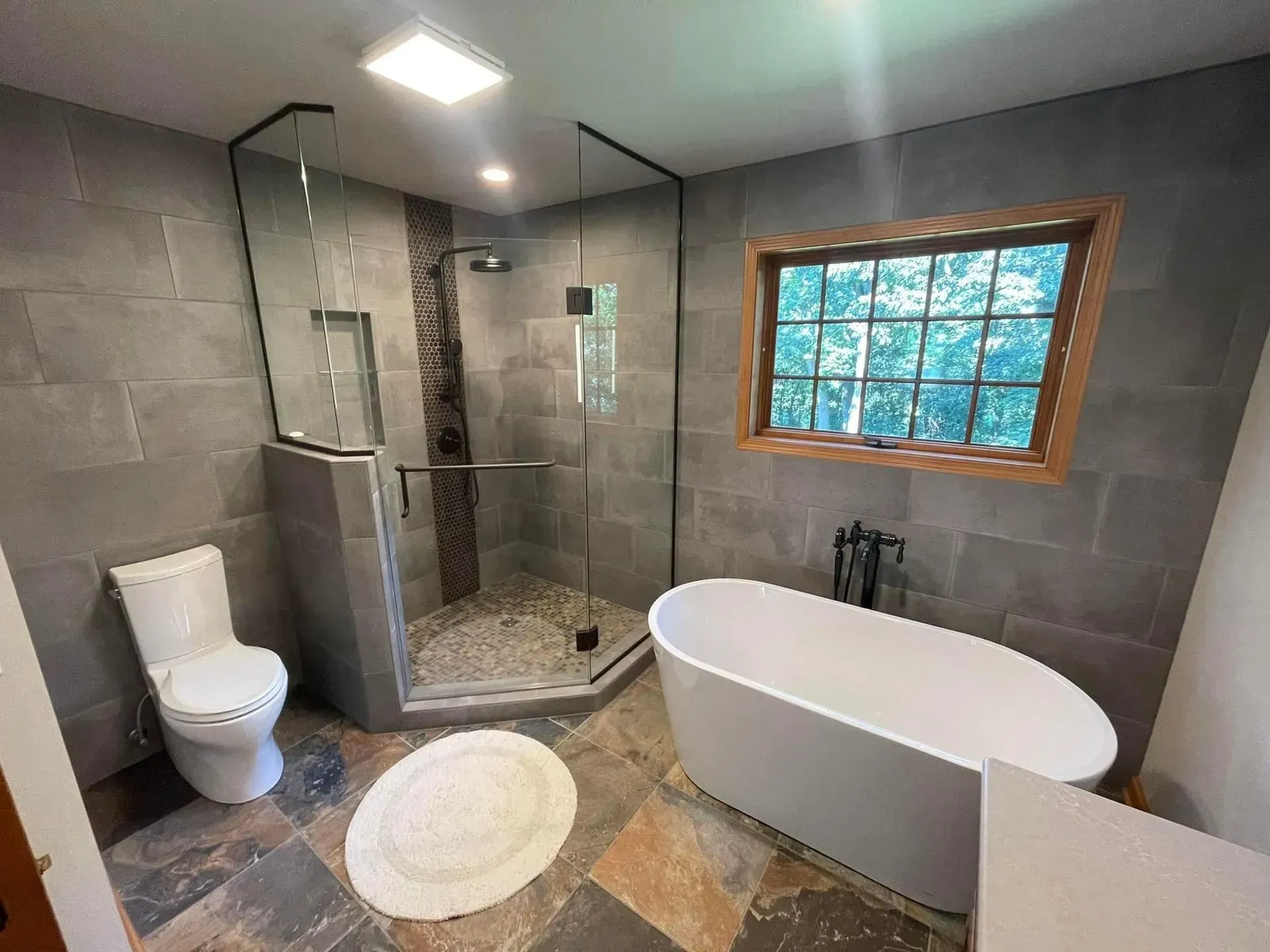 Modern bathroom with gray tile, walk-in shower, freestanding tub, and small window with outdoor view.