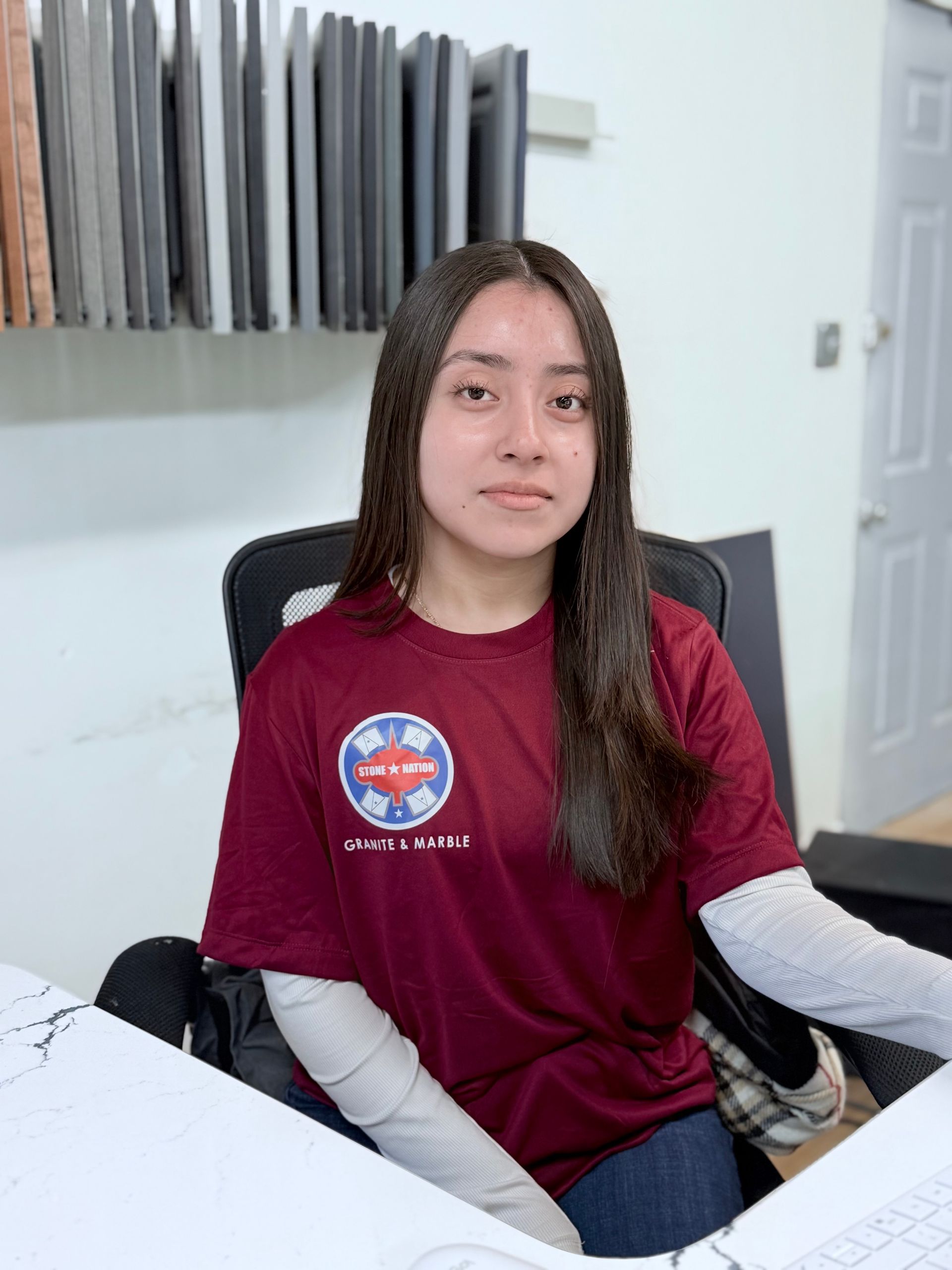 A woman is sitting at a desk wearing a maroon shirt.