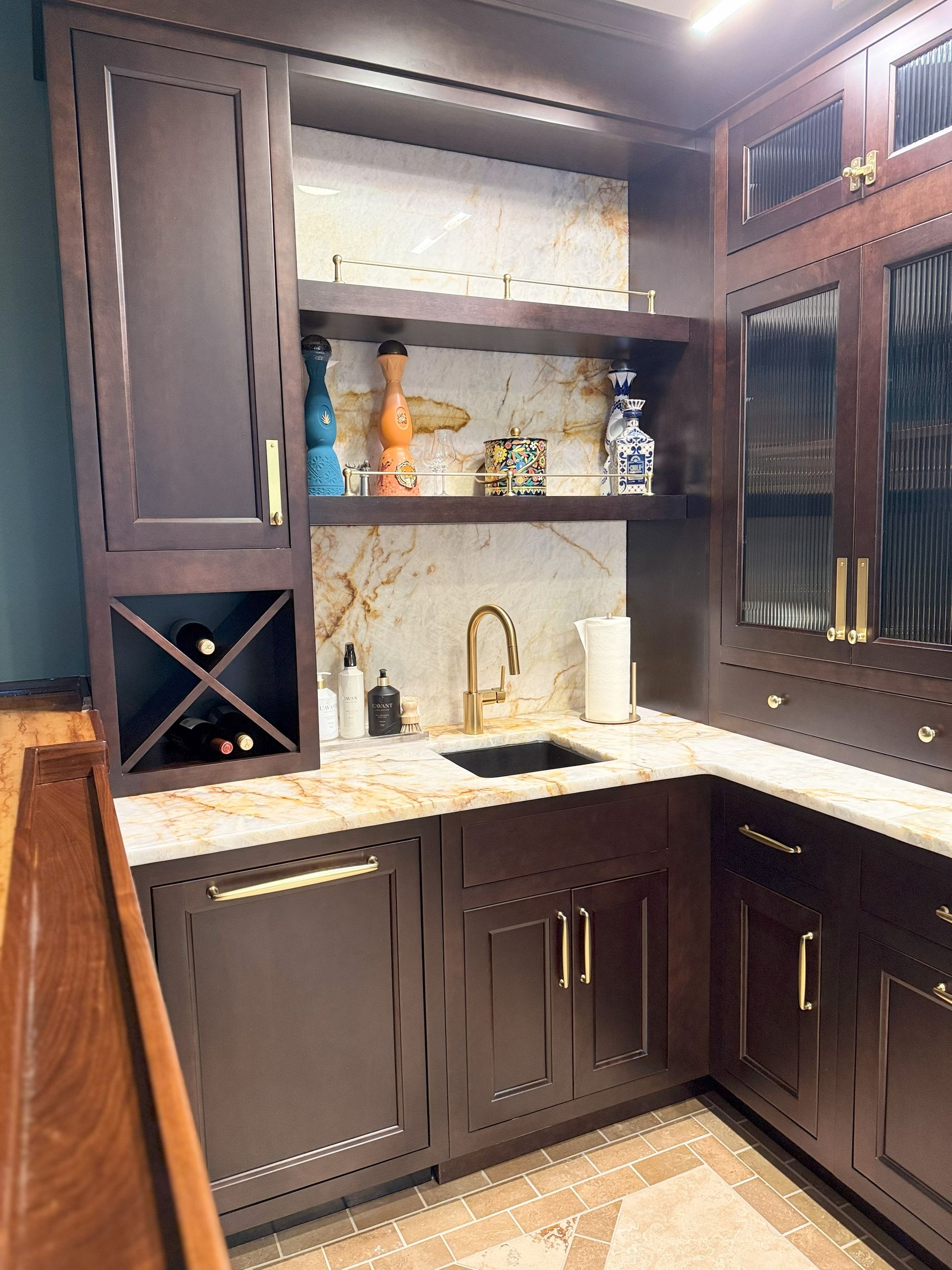Dark wood bar with wine rack, sink, gold hardware, and marble backsplash.