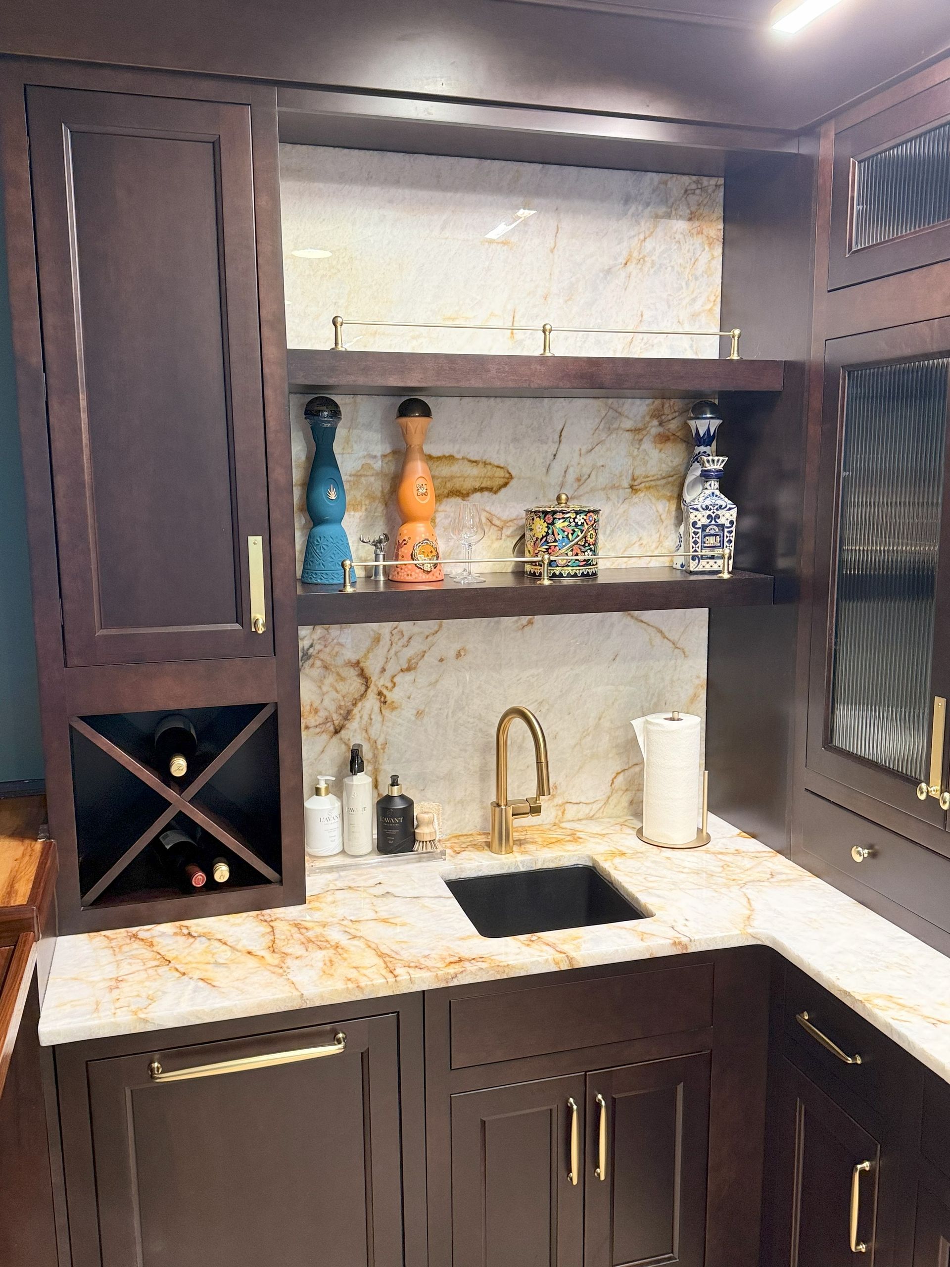 A dark wood home bar with a sink, shelves, and wine storage. Gold accents, marble countertop, and colorful decor.