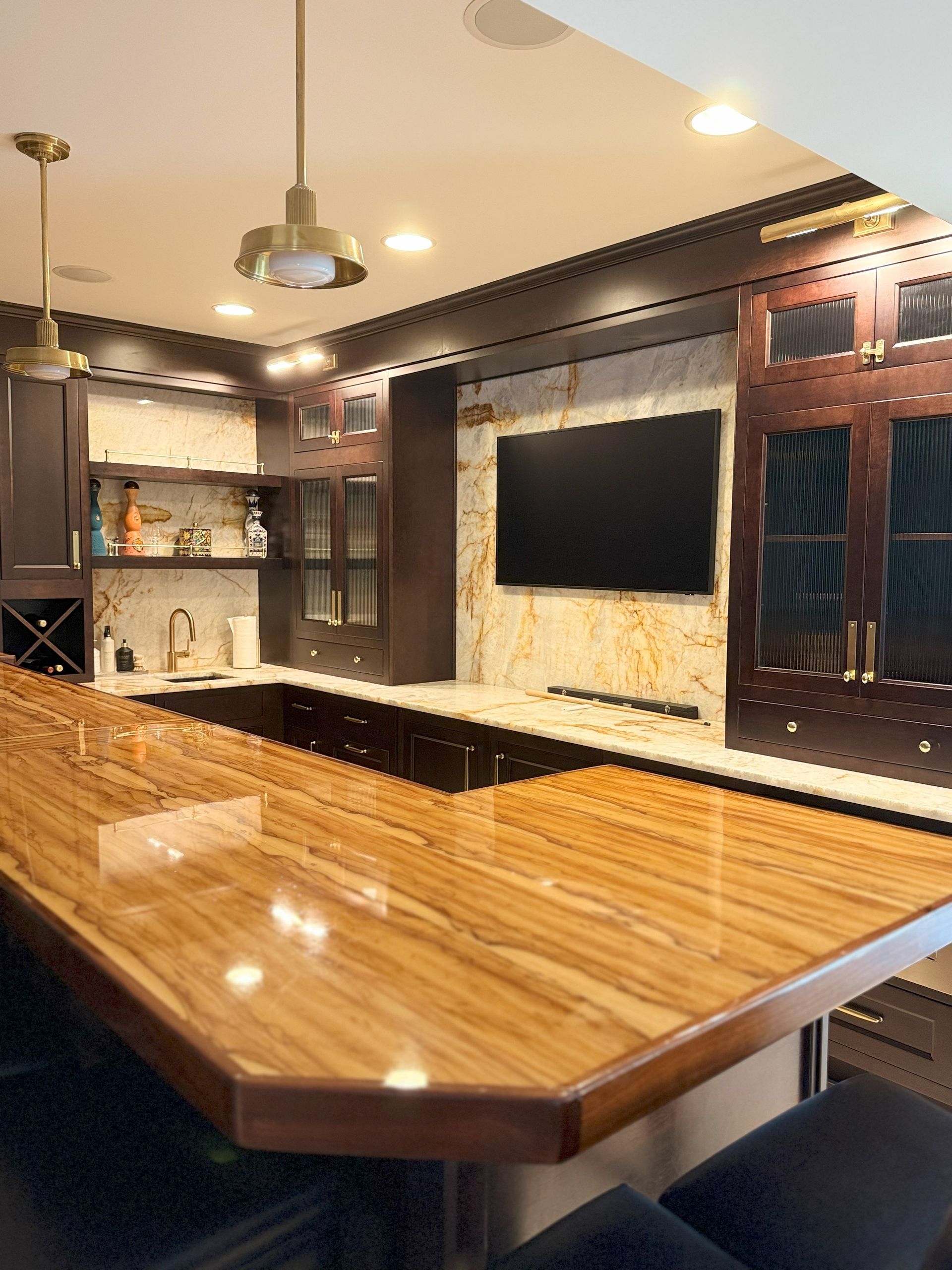 Bar with a light-colored, glossy wood counter, dark wood cabinets, a TV, and pendant lights.