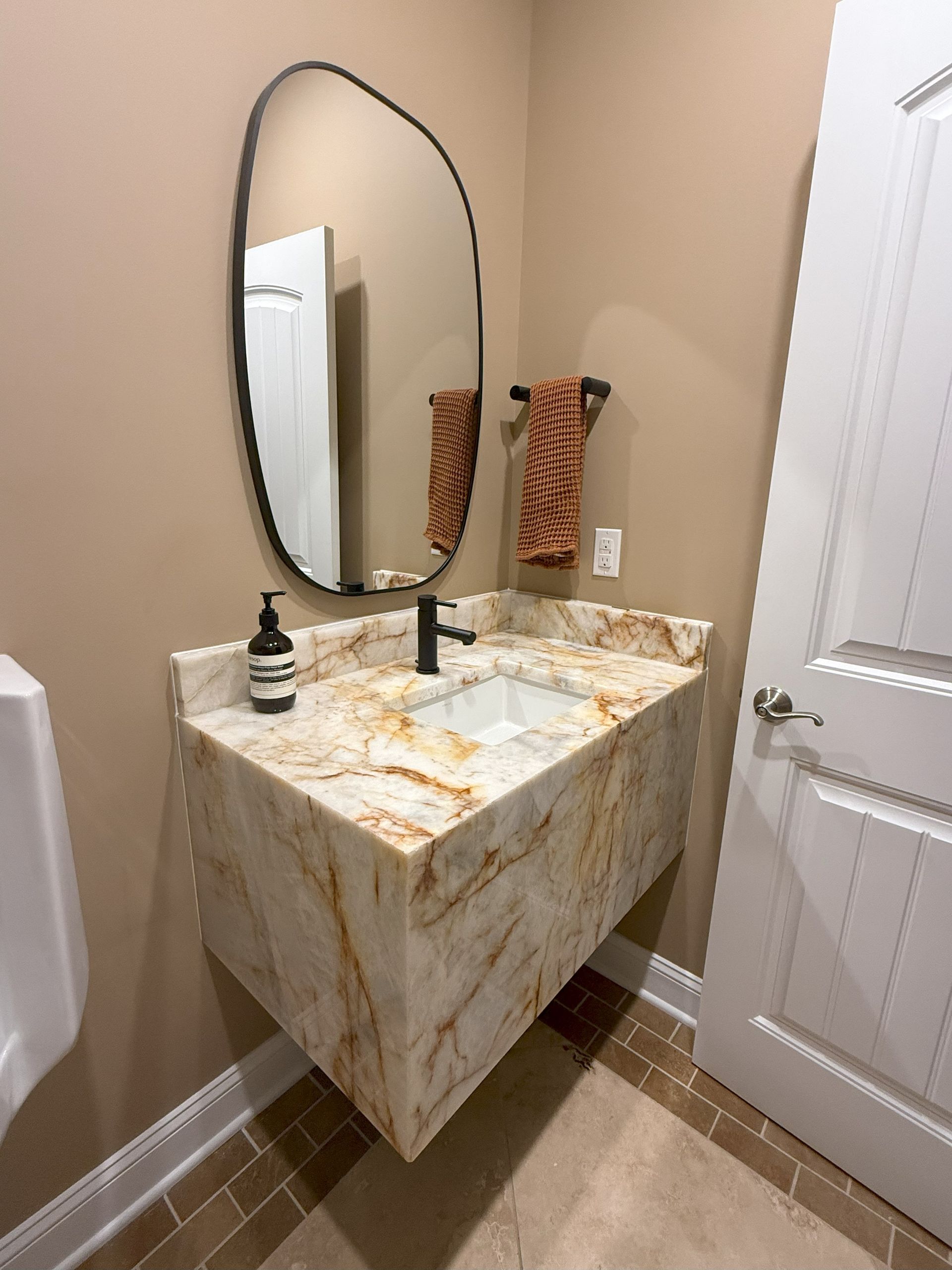 Wall-mounted sink with a marble countertop and freeform mirror in a tan-colored bathroom.