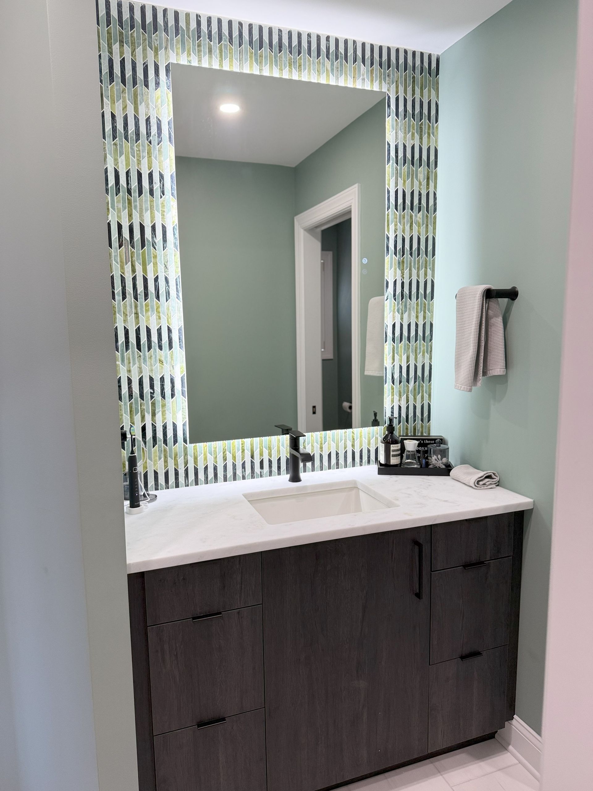 Bathroom with dark gray vanity, patterned mirror frame, light blue-green walls, and black fixtures.