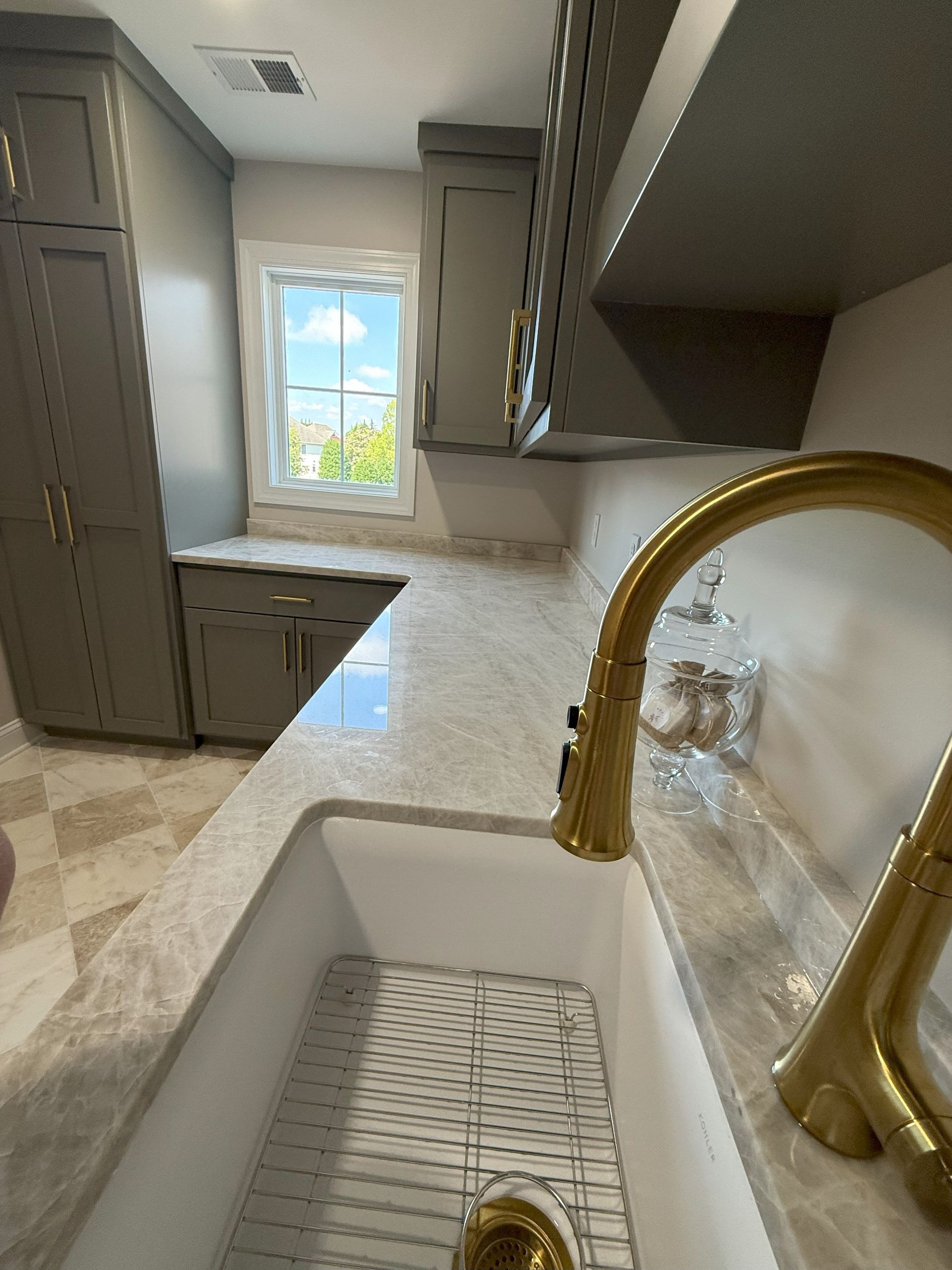 Gold faucet over white farmhouse sink. Kitchen with gray cabinets and light countertops.