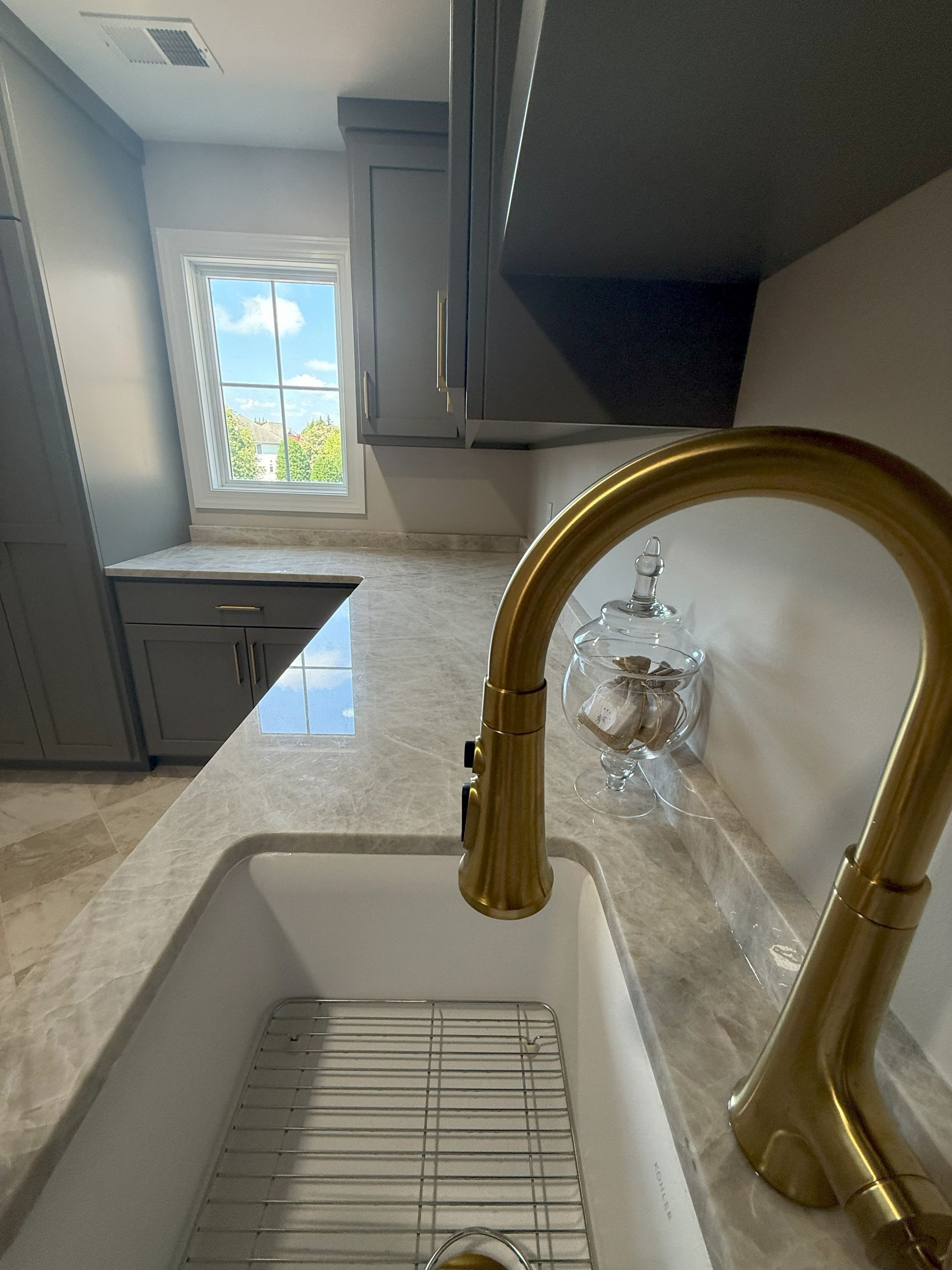 Gold faucet over white kitchen sink; grey cabinets, marble countertop, sunny window.