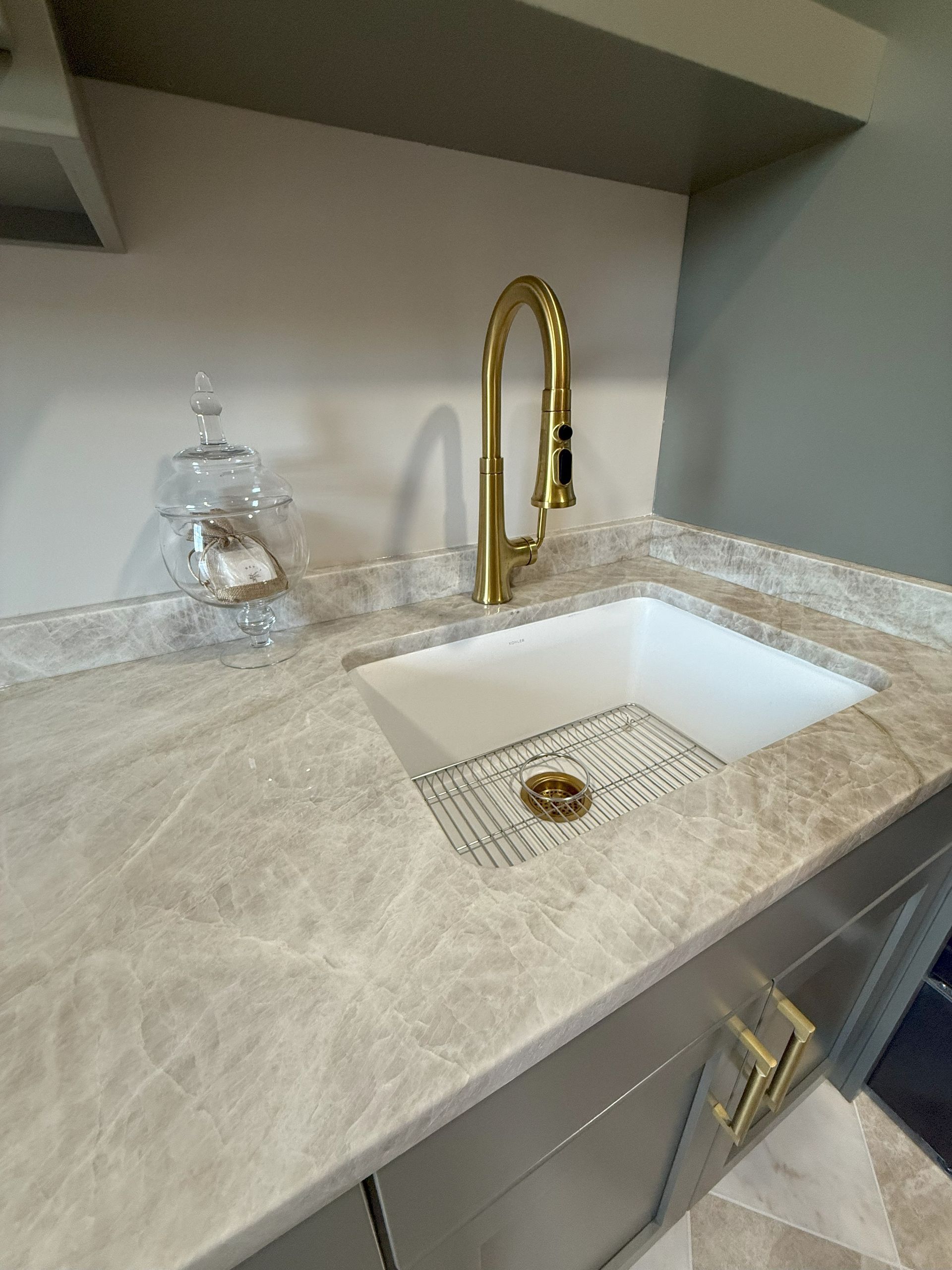 Gold faucet over a white sink with a marble countertop. Grey cabinets with gold handles.