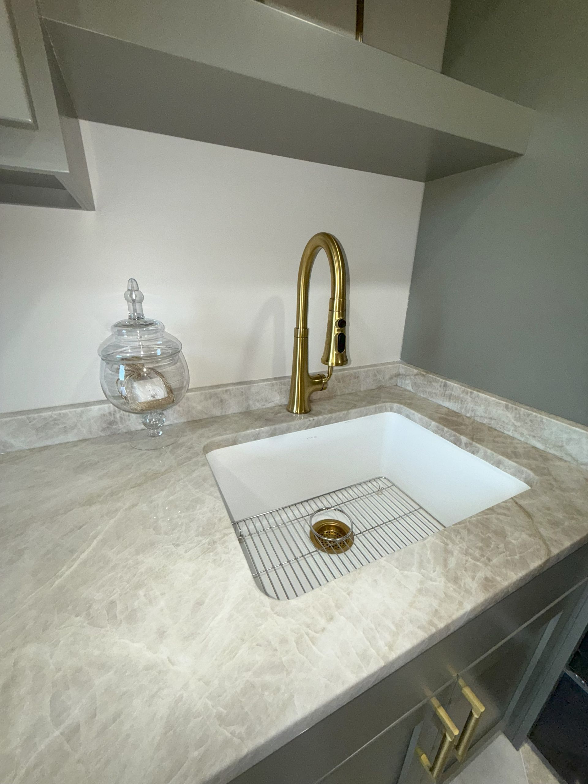 Gold faucet over a white sink on a gray countertop, with a decorative jar and gray cabinets.