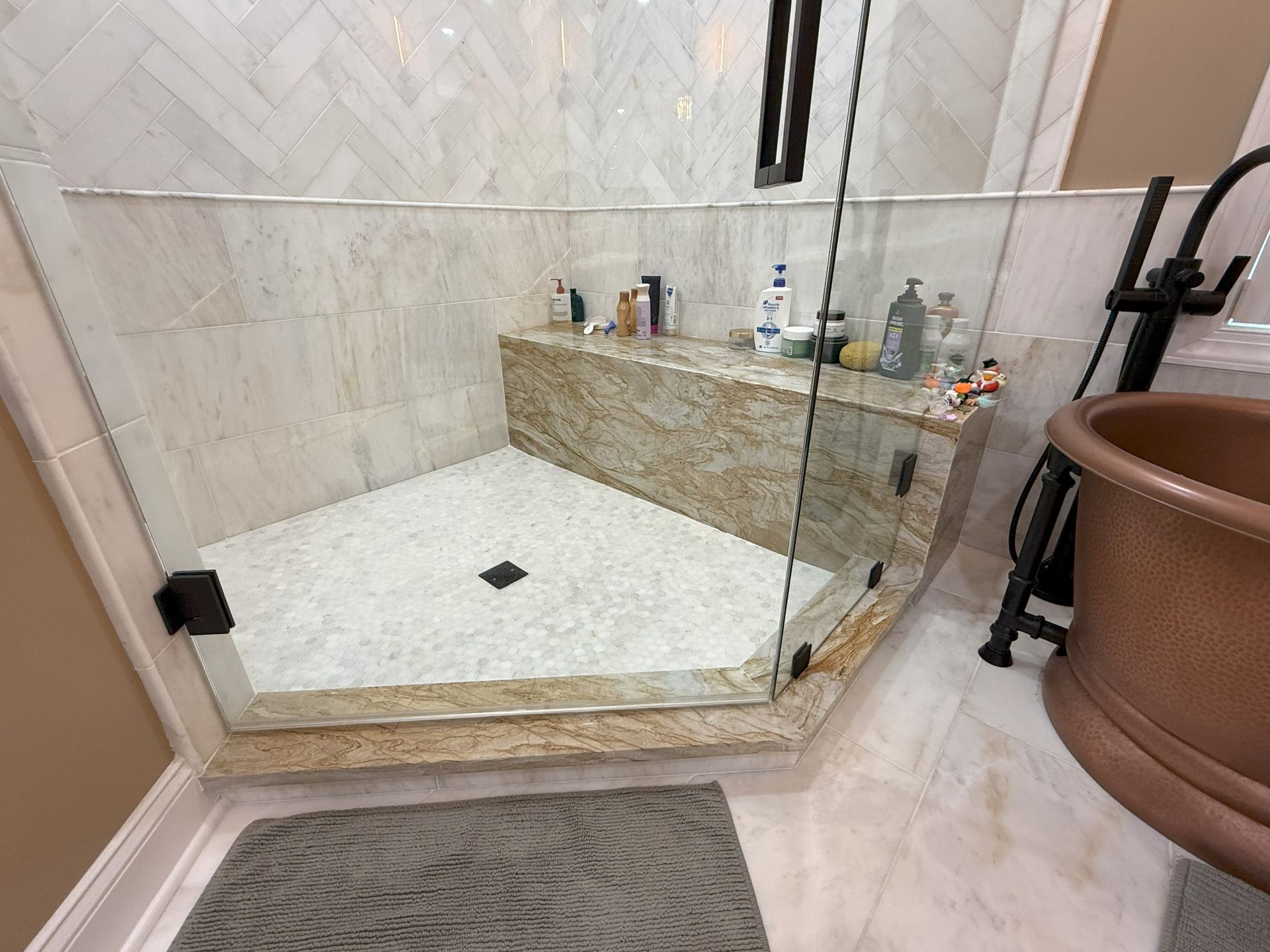 Triangular shower with glass door, beige stone tile, built-in bench, toiletries, and copper tub.