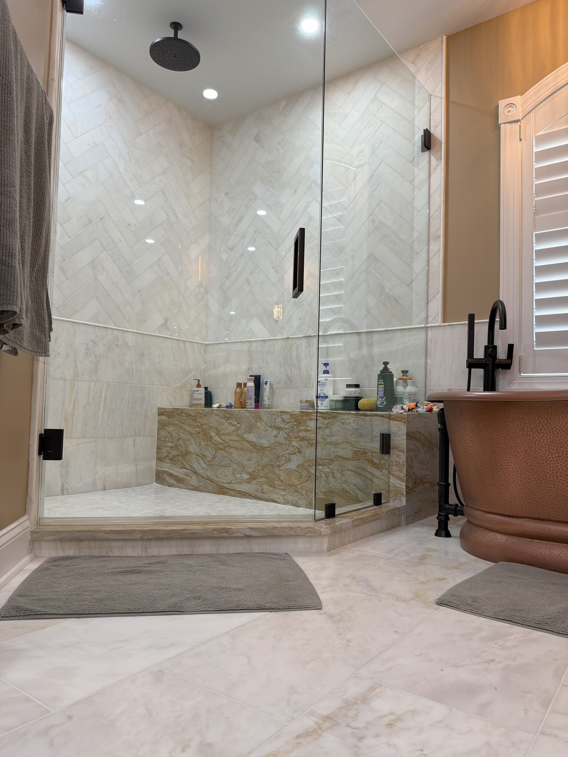 Luxurious bathroom: marble, glass shower, copper tub, tan walls, light gray rug.
