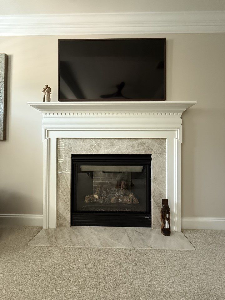 Fireplace with marble tile and white mantel, topped with a TV and decorations, set against a beige wall.