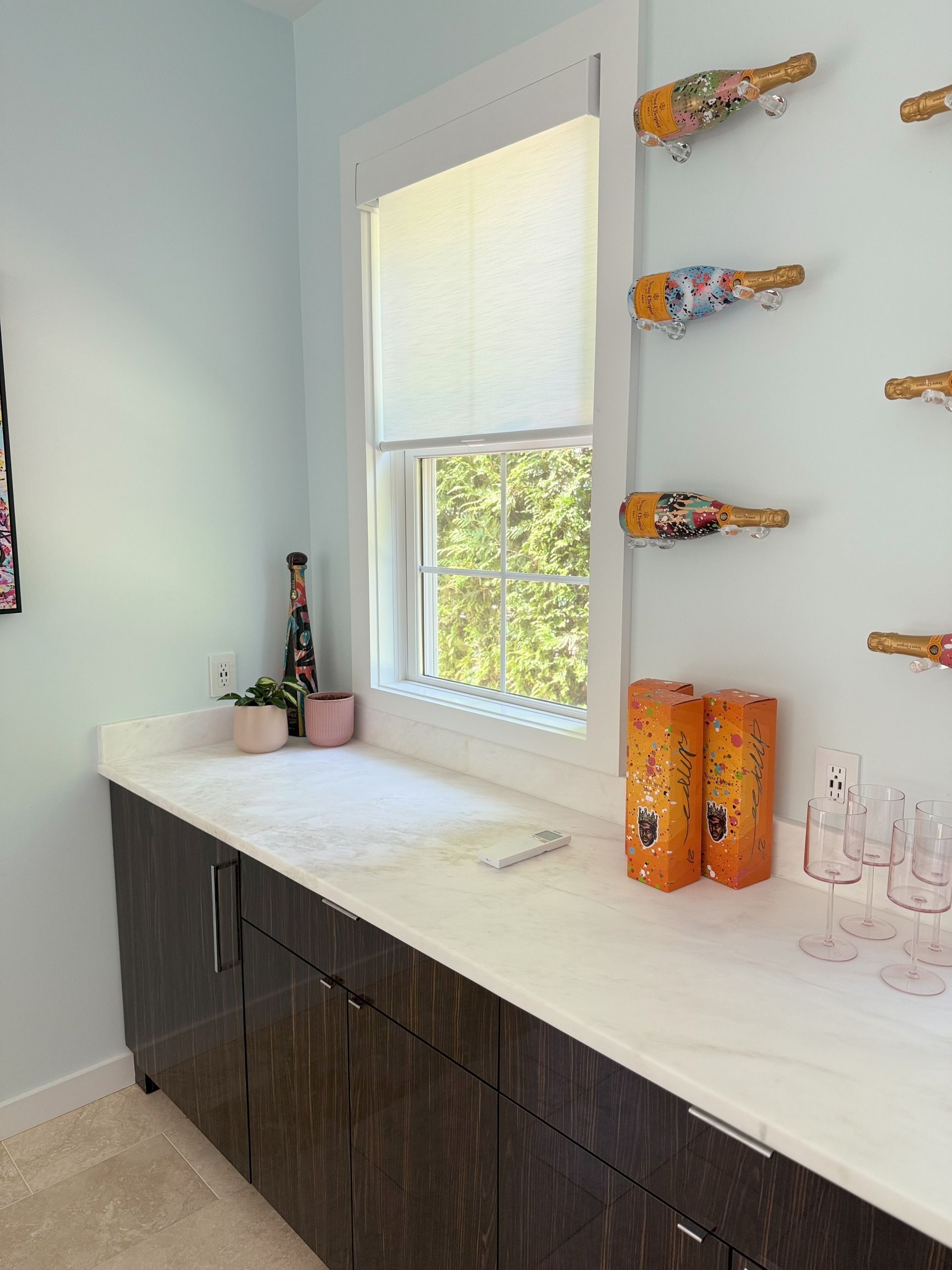 A bar area with a window, cabinets, countertop, wine bottles, and glasses, in a room with light blue walls.