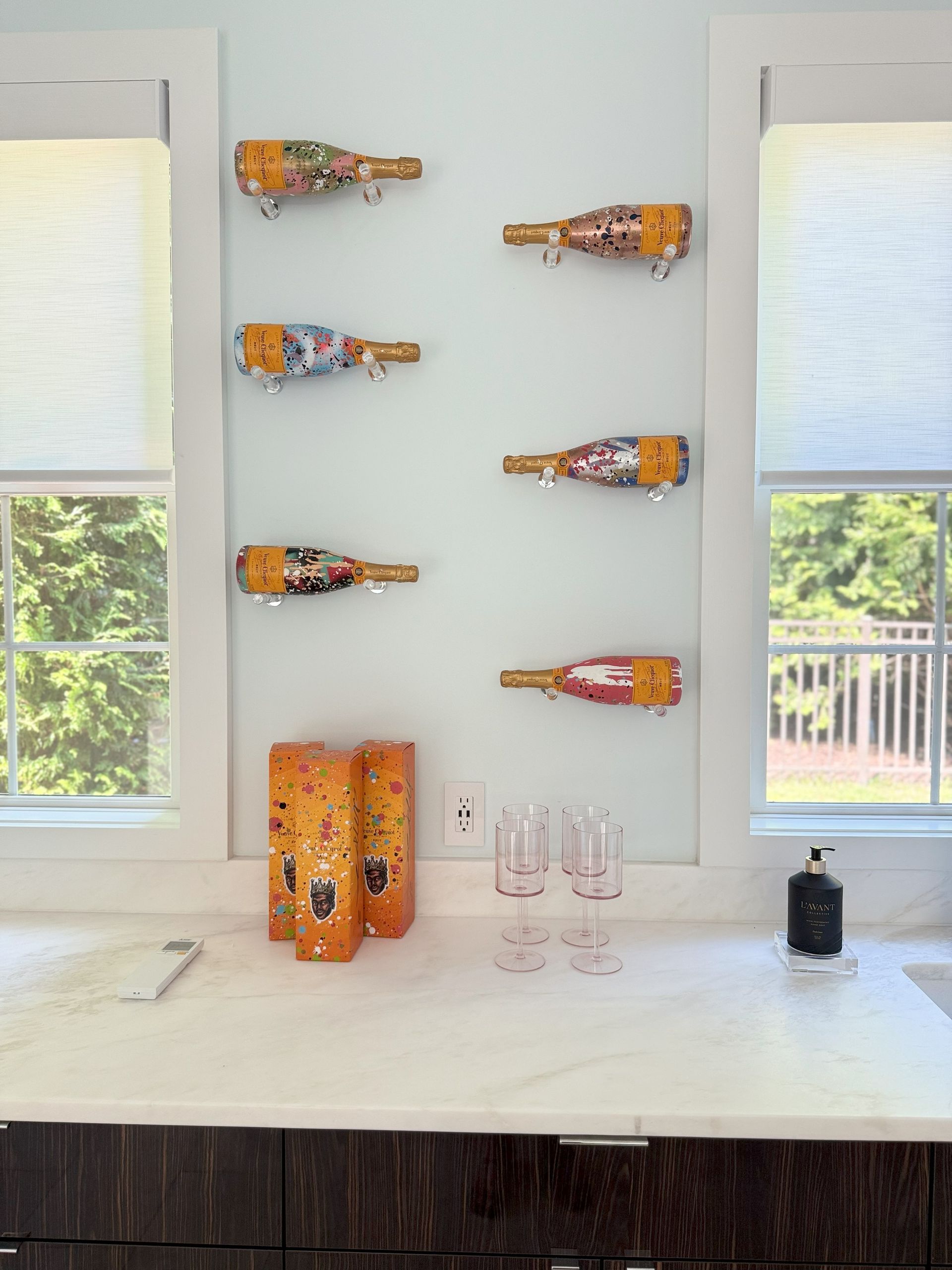 Wine bottle holders mounted on a blue wall above a countertop with glasses, vases, and a window view.