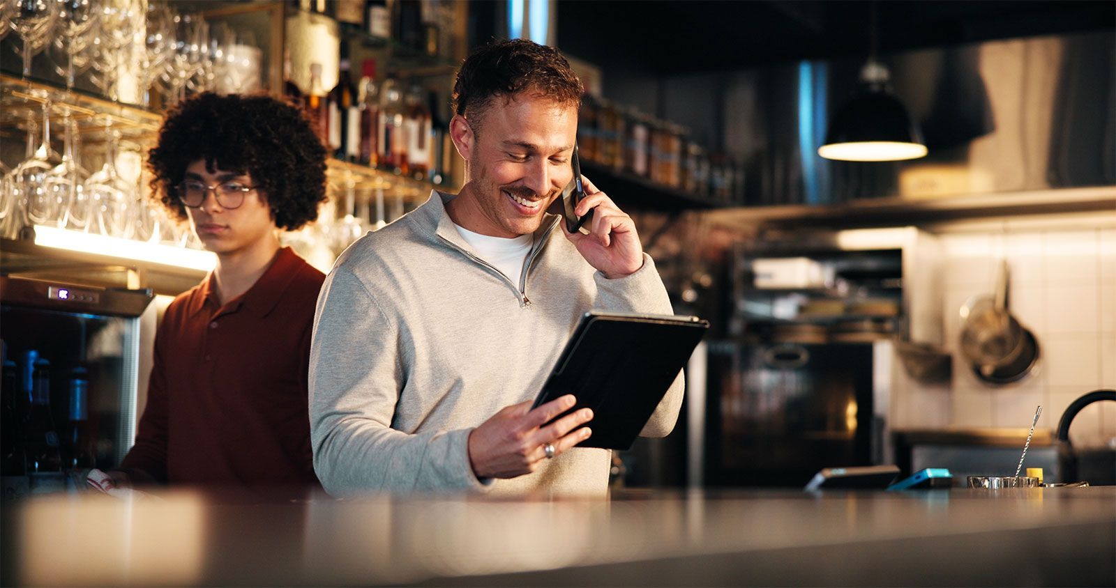 Two people in a warm restaurant kitchen, one smiling while checking a tablet and talking on the phone.