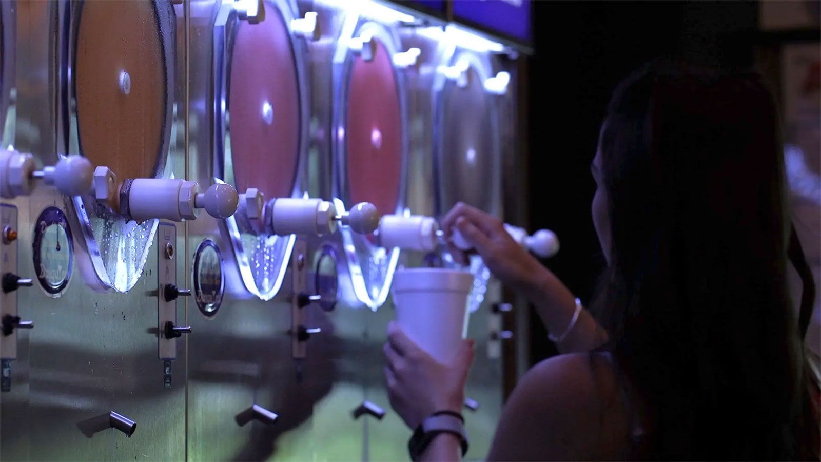 Person filling a cup from a row of frozen yogurt dispensers in a purple-lit shop