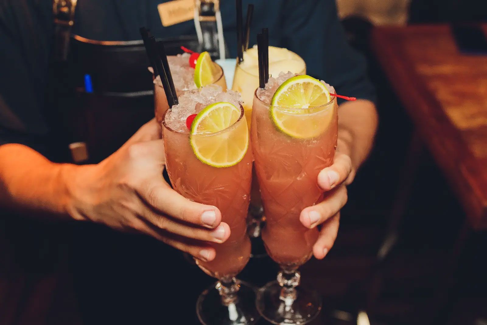 Two hands clinking two orange cocktails with lime and black straws in a dim bar