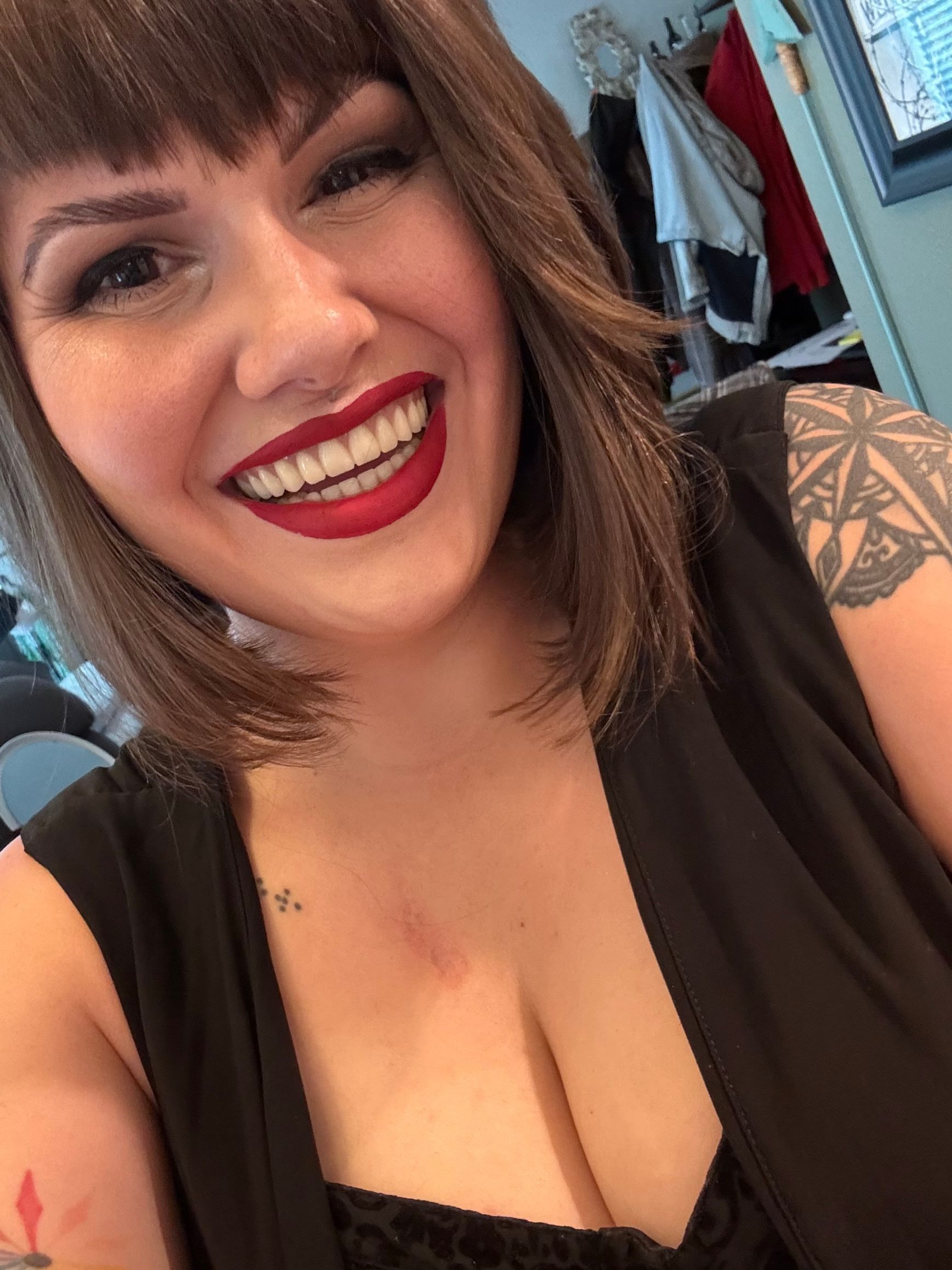Woman with short brown hair, red lipstick, smiling broadly, wearing a black top. Tattoo on arm and small one on chest.