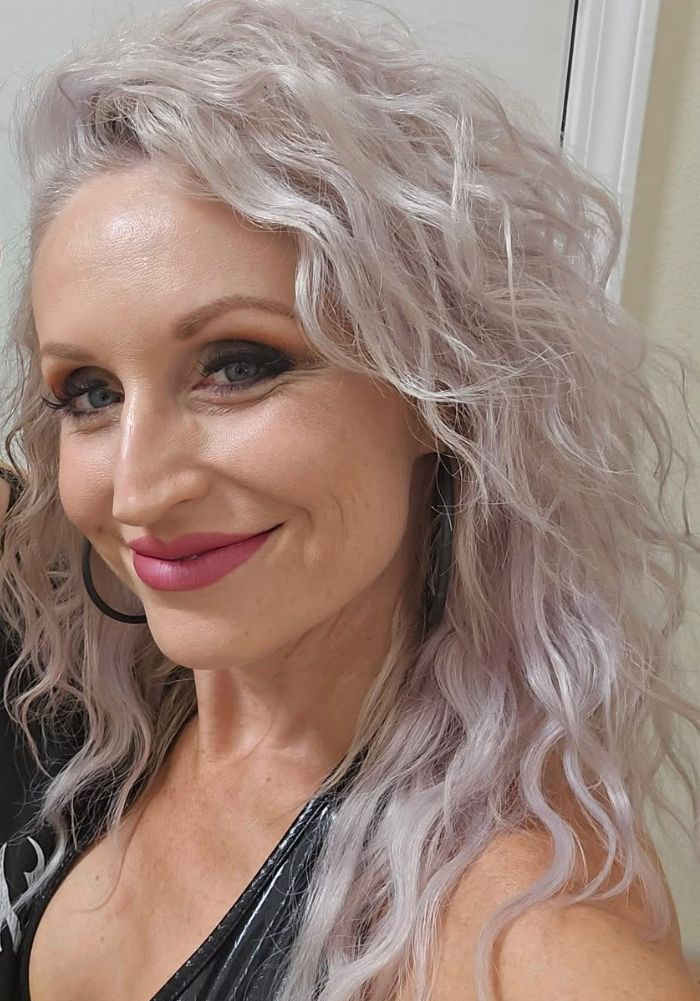 Woman with gray, curly hair smiles, wearing dark eye makeup and pink lipstick.
