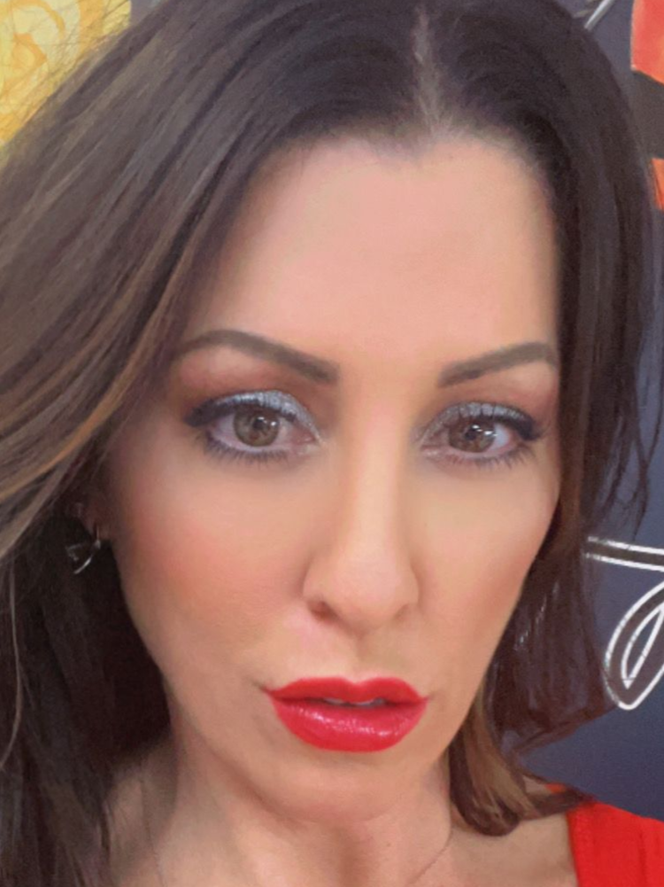 Woman with dark hair, wearing bright eye shadow and red lipstick, looking towards the viewer.