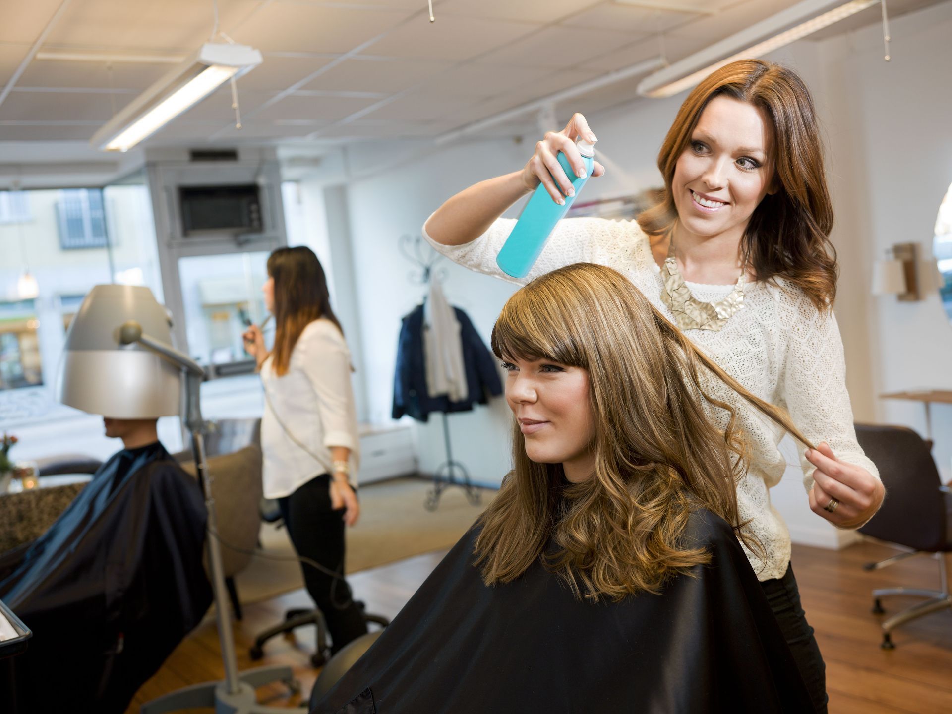 Hairdresser spraying client's hair in salon. Other clients and equipment visible.