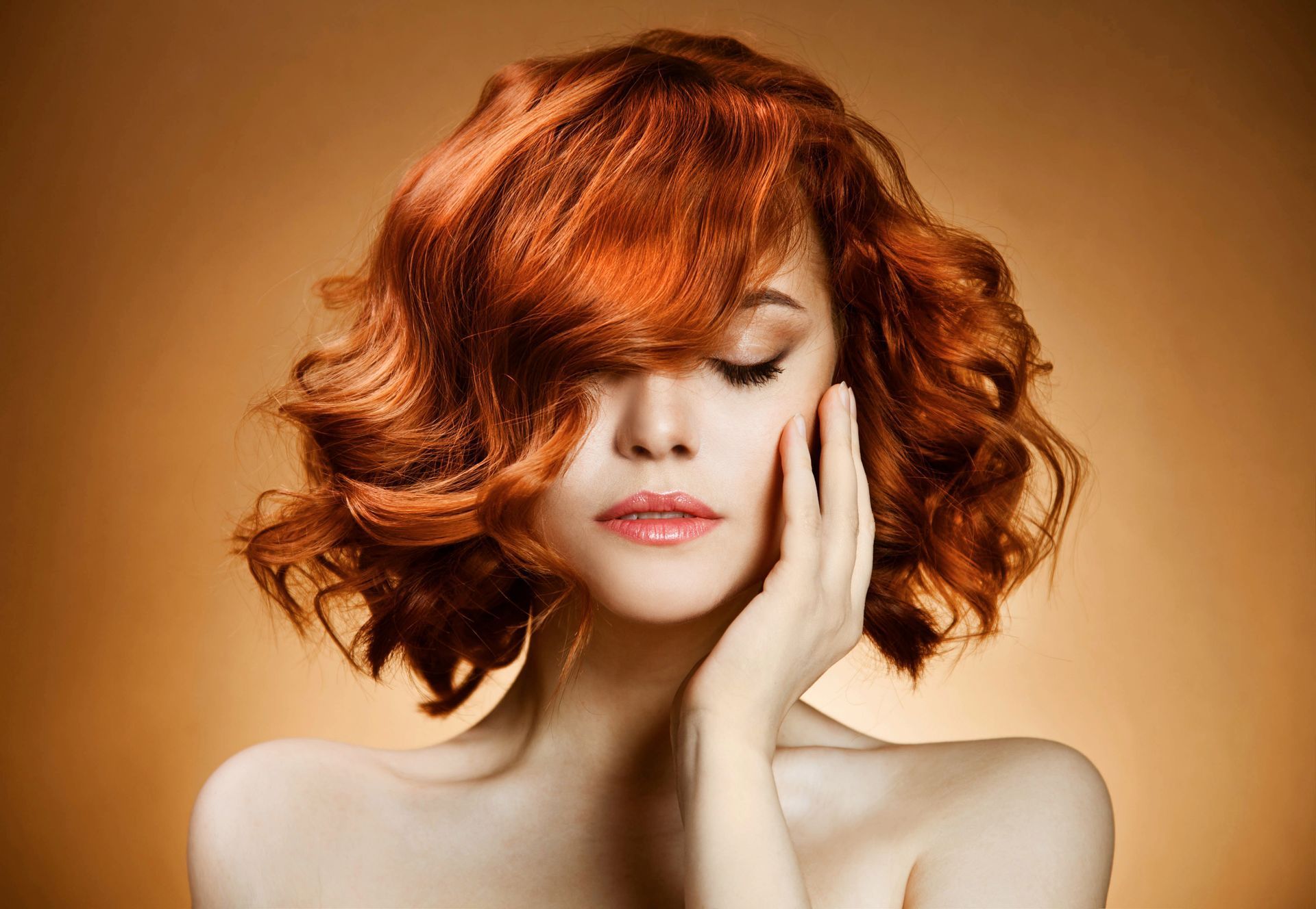 Woman with red, curly hair touching her cheek, eyes closed. Against a golden backdrop.