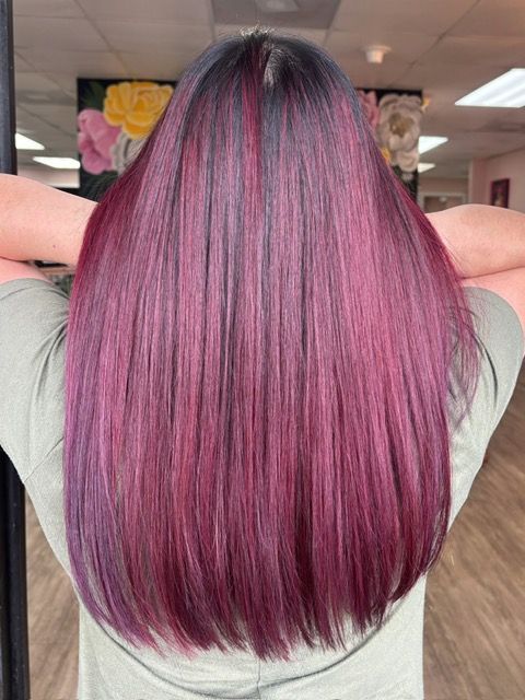 Long, wavy auburn hair down the back against a textured wall, wearing a coral shirt.