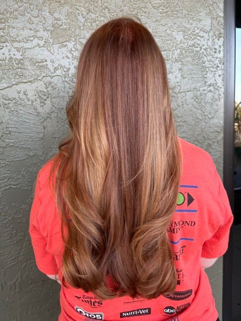 Person with long wavy auburn hair wearing a coral shirt, seen from behind against a textured wall