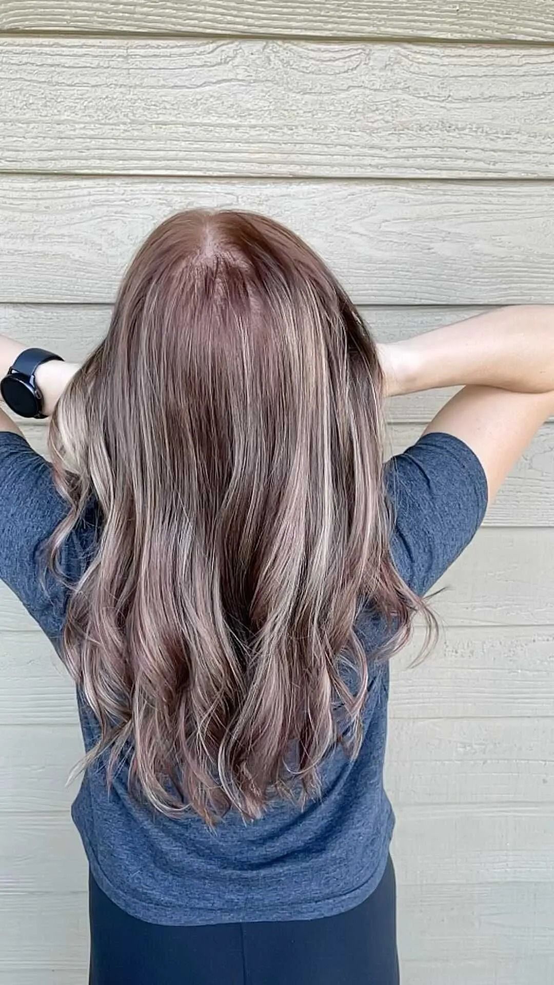 Person seen from behind with hands in hair, wearing a blue shirt against a light wood wall.