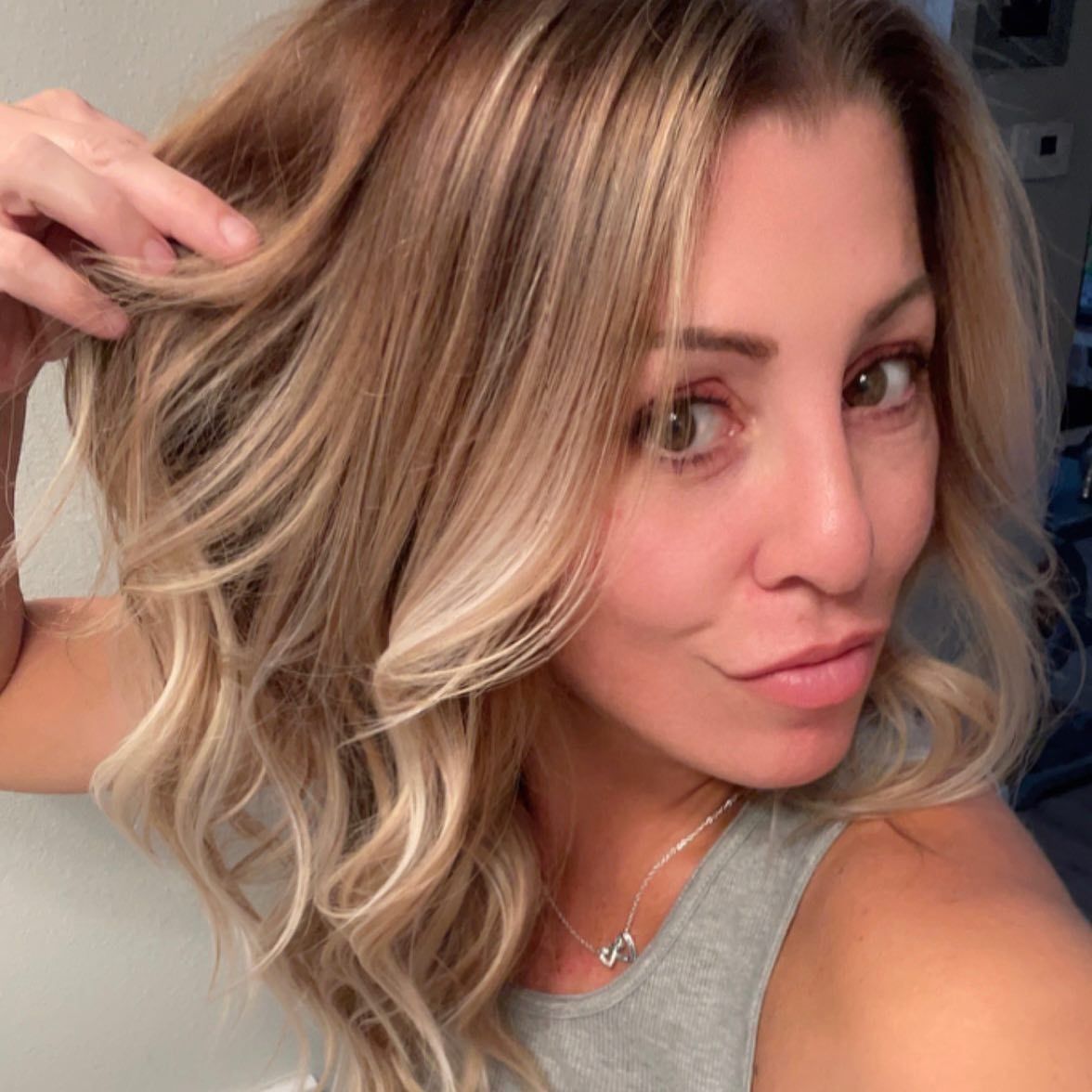 Close-up selfie of a woman with blonde hair, wearing a gray top and necklace, making a playful pout.