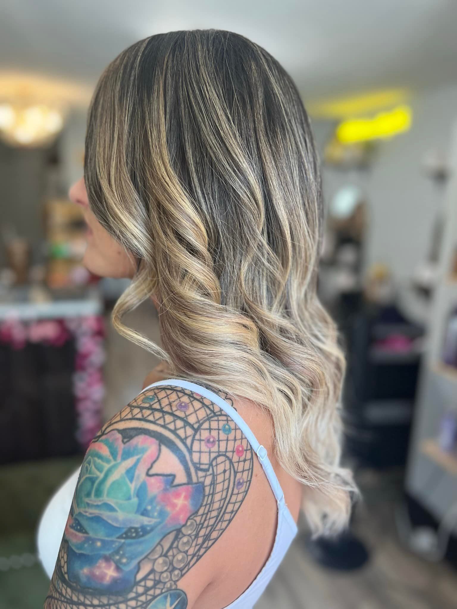 Side-view of a woman with long blonde hair and a colorful tattoo, standing in a salon.