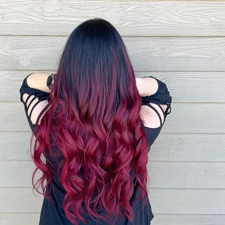 Woman with long, wavy black hair that fades into vibrant red. Leaning against a wood wall.