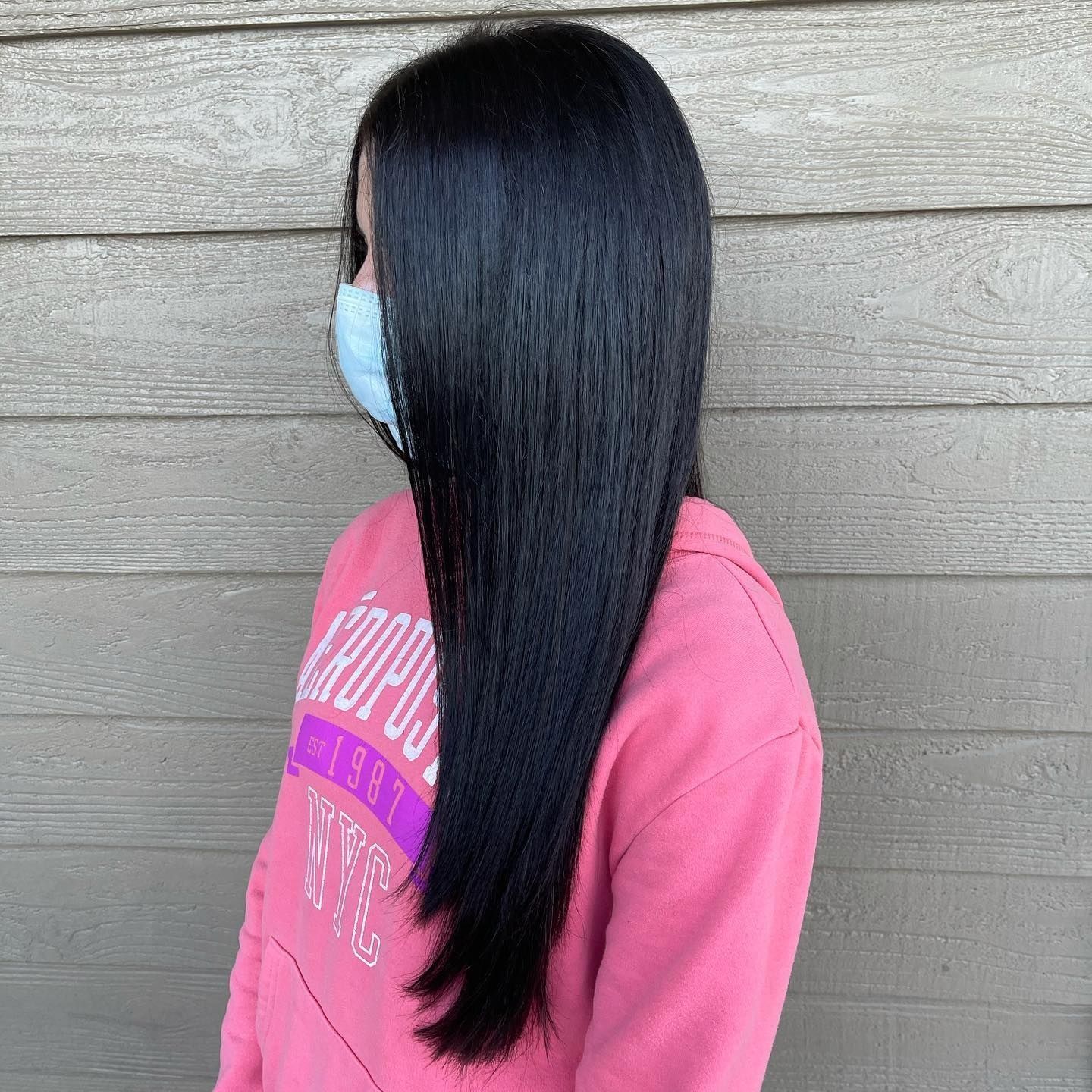Person with straight, black hair, wearing a light blue mask and pink sweatshirt in front of a wood-paneled wall.