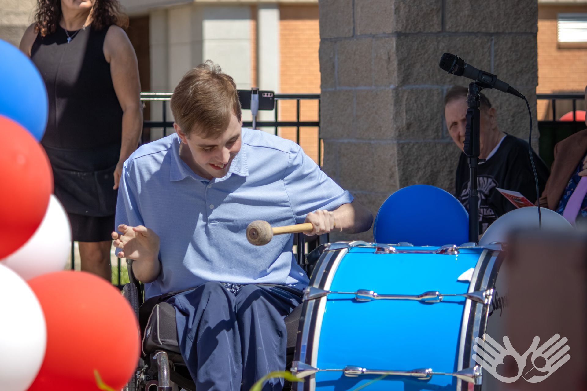 Boy playing a blue drum outdoors at a performance, with balloons and people nearby