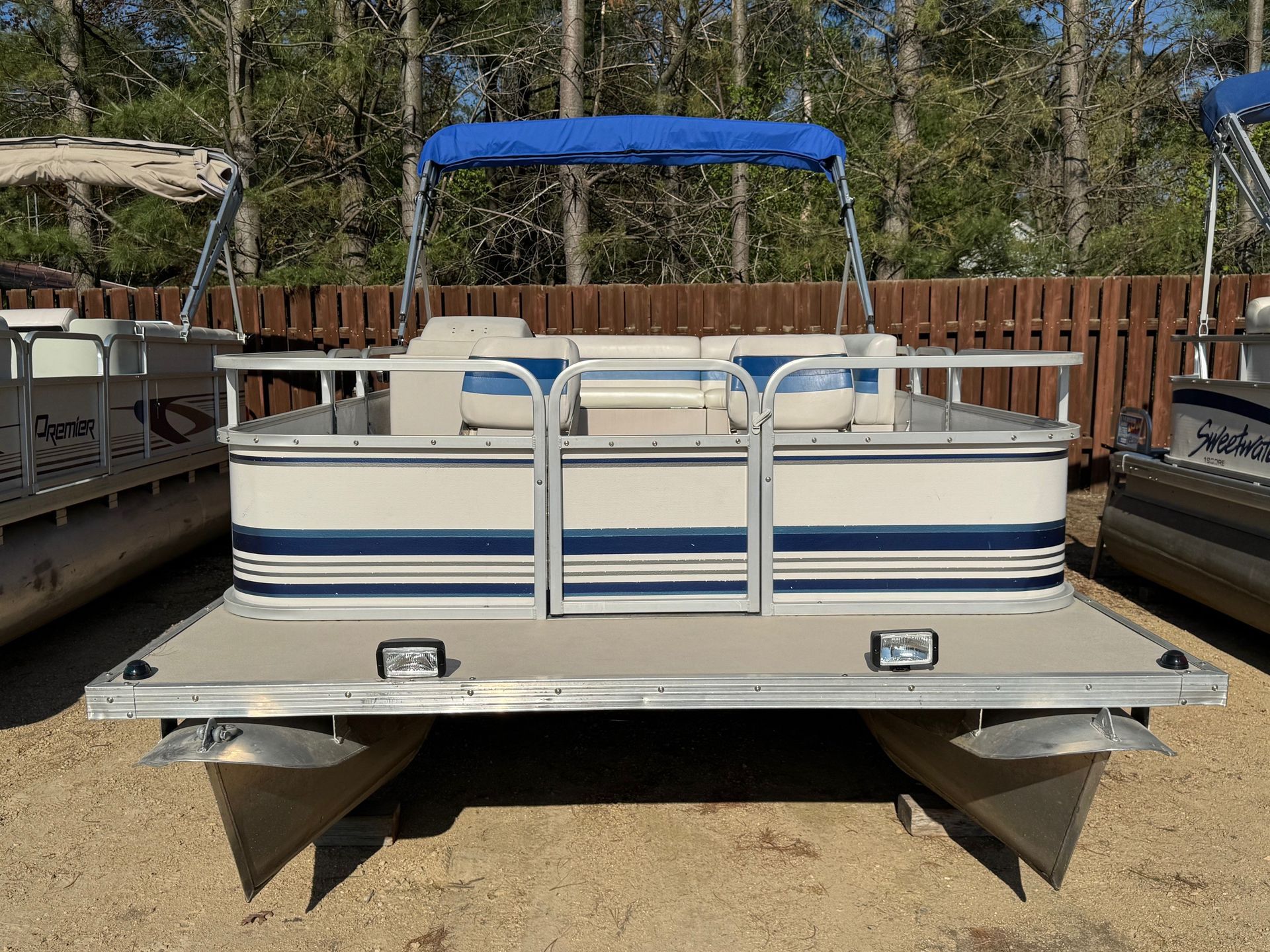 A pontoon boat with a blue canopy is parked in front of a wooden fence.