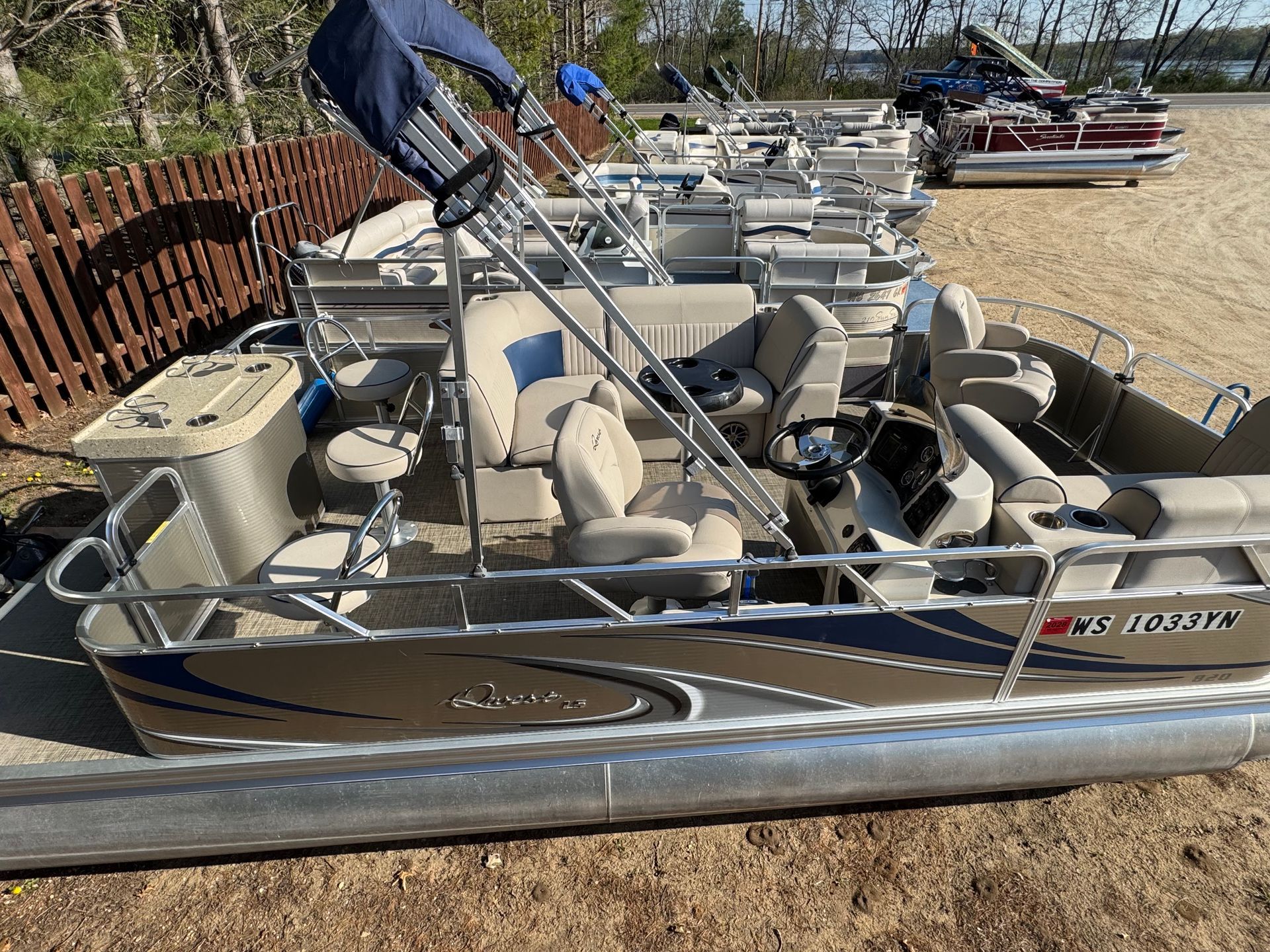 A pontoon boat is parked in a lot next to a fence.