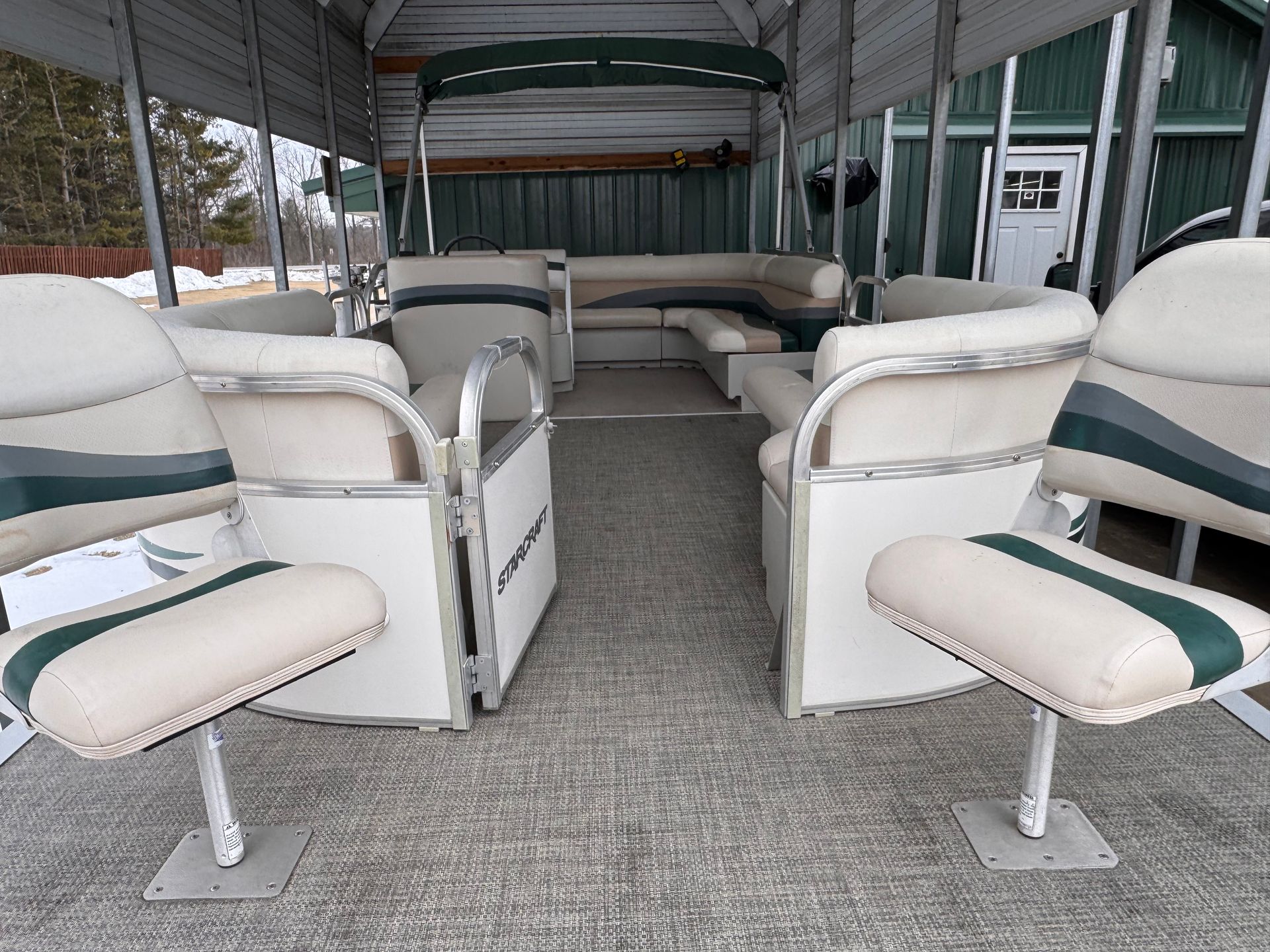 Interior of a pontoon boat featuring cream and green upholstered seating under a canvas canopy inside a storage building.