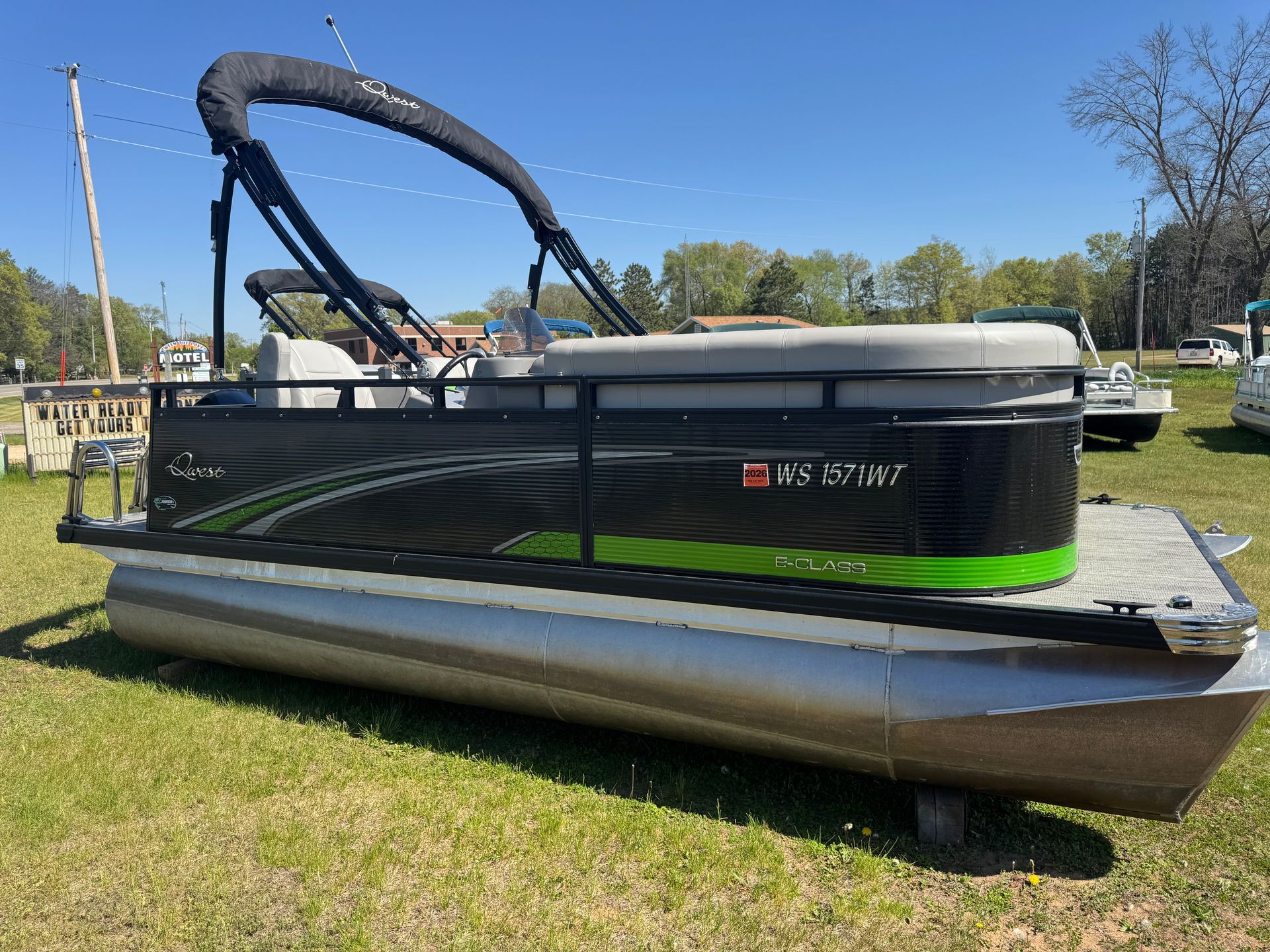 A beige and green pontoon boat with a dual-canopy top parked on a gravel lot near a wooded area.