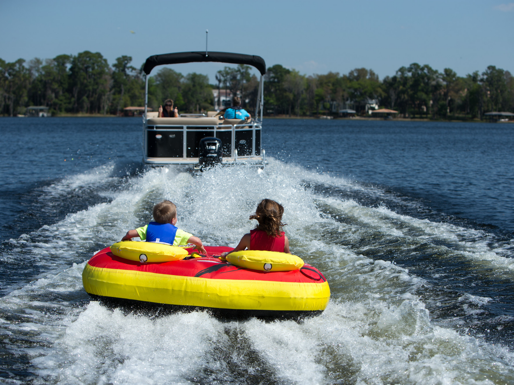 A pontoon boat is floating on top of a body of water. Kids tubing behind pontoon.